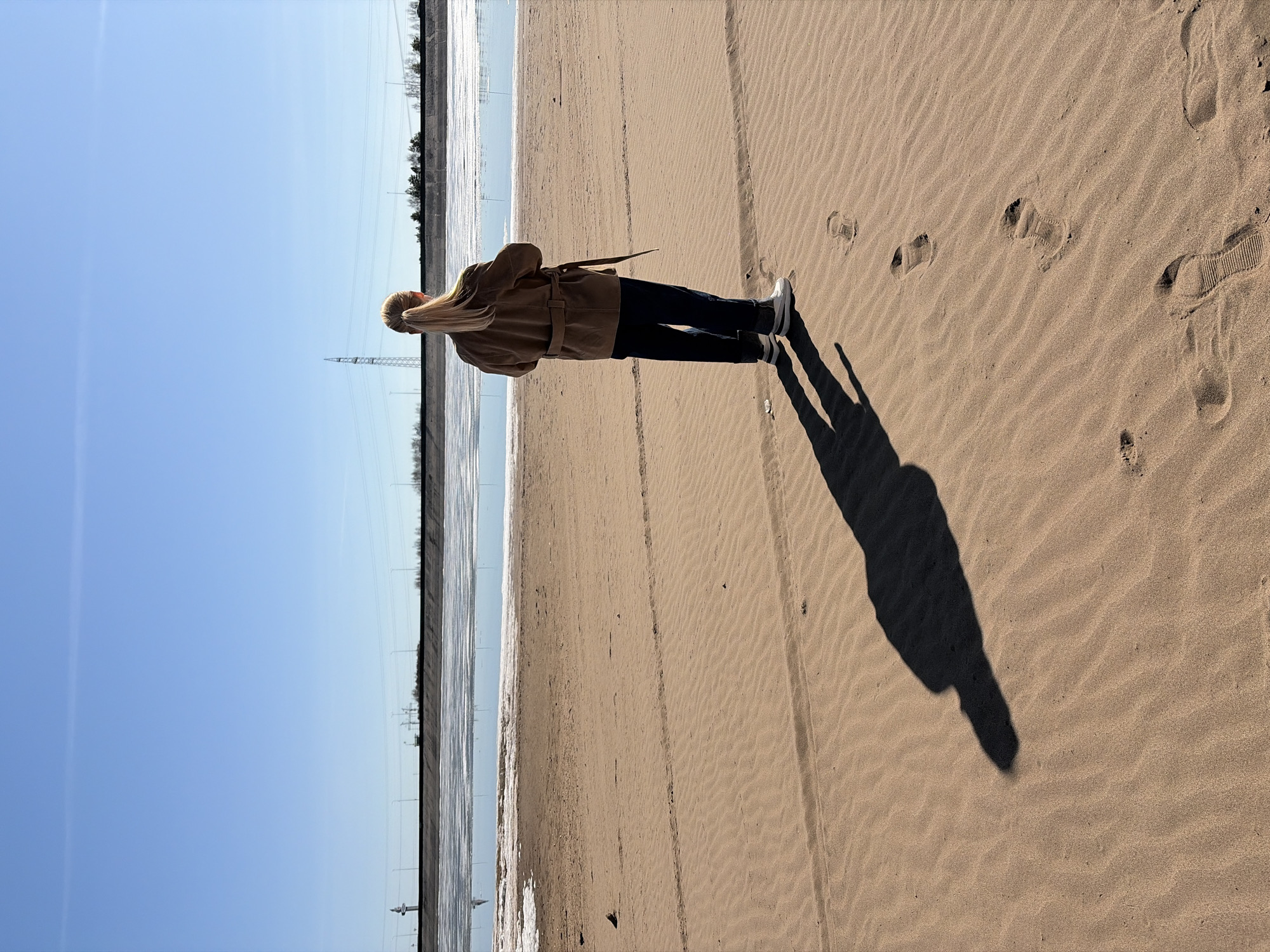 The image, oriented correctly, depicts a person standing on a sandy expanse, gazing towards a body of water under a clear blue sky. The individual, seen from behind, has long blonde hair tied in a ponytail and is wearing a light brown or tan belted jacket, dark trousers, and white and black sneakers. A long, dark shadow of the person stretches across the textured sand, indicating a low sun angle, likely early morning or late afternoon.
The setting appears to be a wide beach or riverbank. The sandy ground shows subtle rippled patterns and several footprints, suggesting recent movement. Beyond the sand, a body of water, possibly partially frozen or covered in ice, extends across the mid-ground. In the distance, power lines and tall power line towers are visible, running parallel to the water's edge, with a line of sparse trees or vegetation on the far horizon. The bright, cloudless sky suggests fair weather. There is no visible text in the image. The scene is likely captured in Noviy, Russia, providing context to the landscape and conditions.