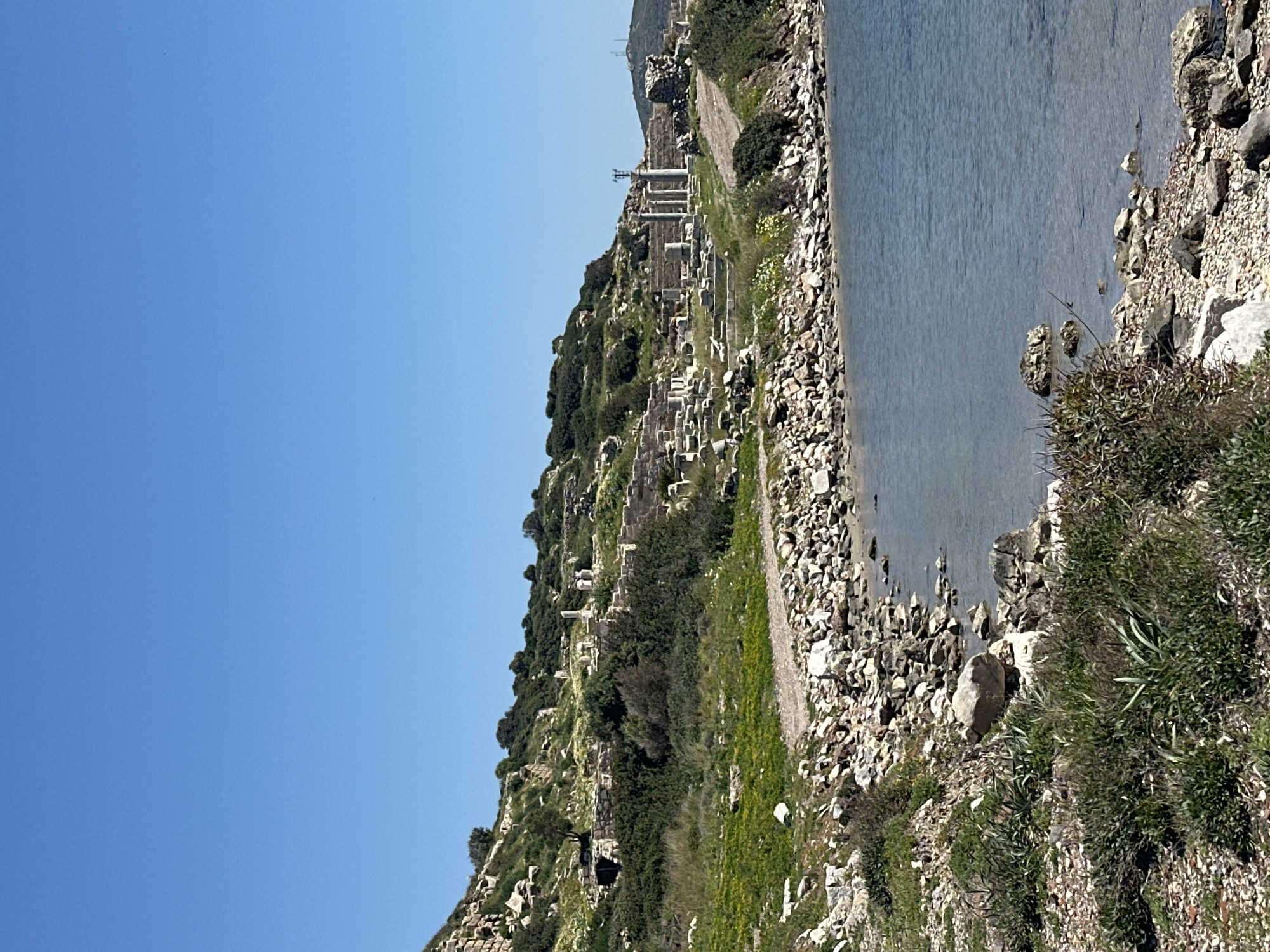 The image shows the ancient city of Knidos near Datça, Turkey, situated on a hillside overlooking the sea. The site is characterized by ruined stone structures, including pillars, and remnants of walls scattered across the slope. The hillside is covered with sparse vegetation, a mix of grass and shrubs. The shoreline consists of rocks and pebbles, and the sea appears calm. A clear blue sky dominates the upper left portion of the image. There are no people visible in the scene.