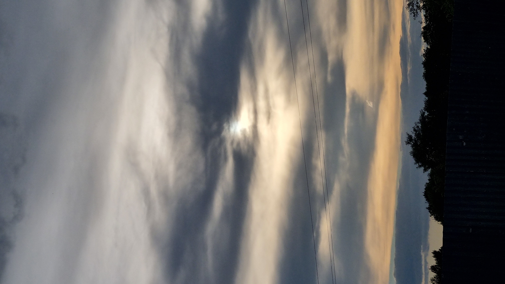 The image is a vertical, low-angle outdoor photograph of a cloudy sky with a small portion of the sun visible through a break in the clouds. The sun appears as a bright, diffused light source. The clouds are varied in color, ranging from dark gray to lighter grays and pale yellow, suggesting the time of day is likely dusk or dawn. Three horizontal wires are visible across the mid-section of the frame. In the lower portion of the image, a dark silhouette of trees and a fence can be seen against the lighter sky. The image is taken from a low vantage point looking upwards towards the sky. There are no people or animals visible in the image. The environment appears to be an outdoor setting, possibly in a rural or suburban area given the presence of trees and a fence, and the wire infrastructure. The specific location of Vysokoe, Russia is not discernible from the visual information presented in the image.