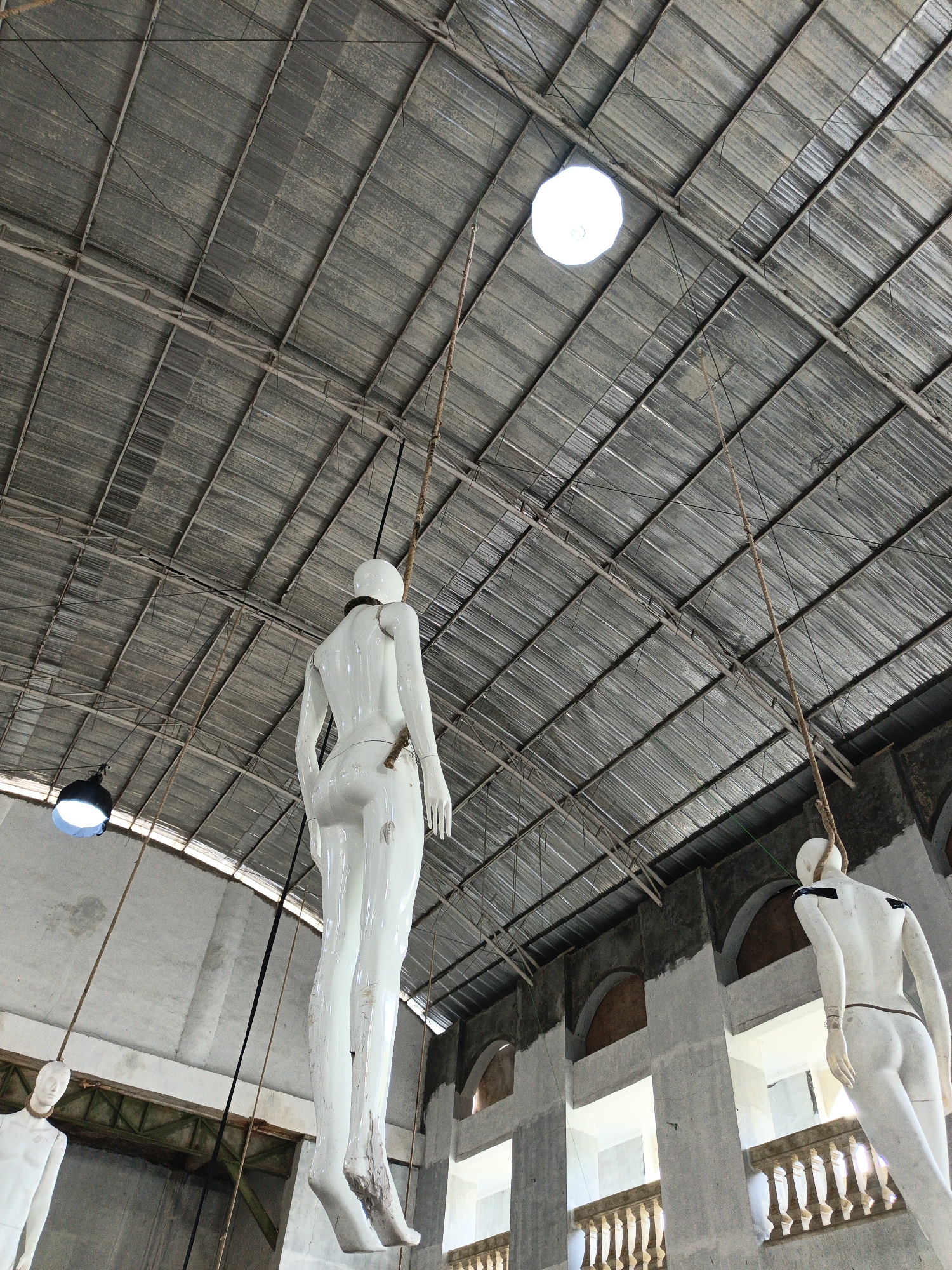 The image depicts an indoor scene with a high, corrugated metal roof supported by a network of metal beams. Several white mannequins are suspended from ropes attached to the ceiling. One mannequin, in the foreground, is viewed from behind and below, showing its full body hanging. Another mannequin is partially visible on the left, and a third is on the right side of the frame, also hanging. Two industrial-style light fixtures are visible: one large, circular light fixture hanging from the ceiling in the upper center, and a smaller, cone-shaped fixture on the left. The walls of the structure appear to be concrete, with arched openings and decorative railings visible in the background. The overall impression is of an abandoned or disused industrial space. The location is specified as Cibogo, Indonesia.