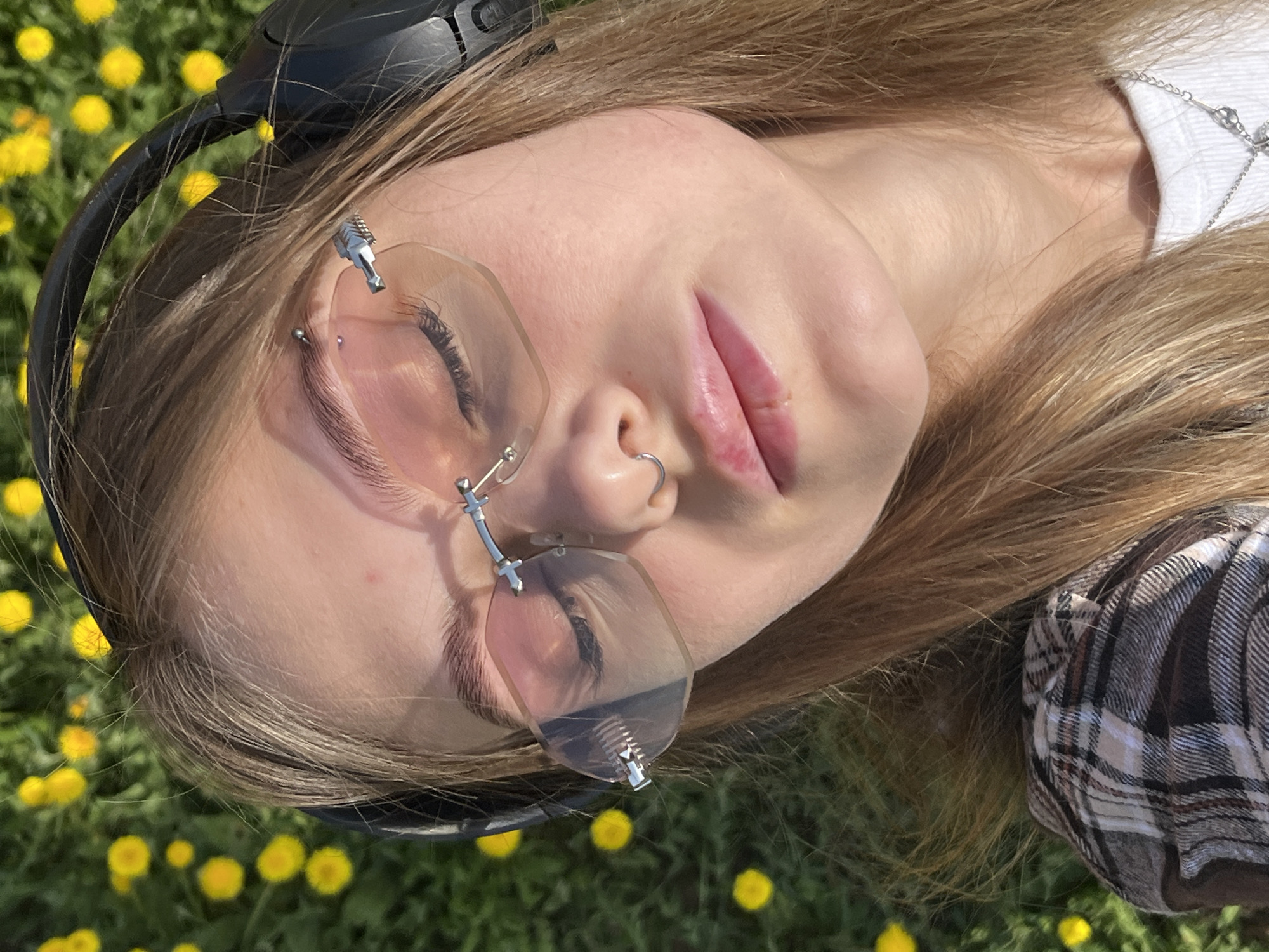 A young woman is lying on her back in a grassy field dotted with yellow flowers. She is wearing headphones and unusual clear-framed glasses. A nose ring is visible. Her hair is light brown, and she's wearing a plaid shirt and a white undershirt with a silver chain. The image appears to be taken outdoors in natural light. The location is not identifiable as Фоки, Russia.