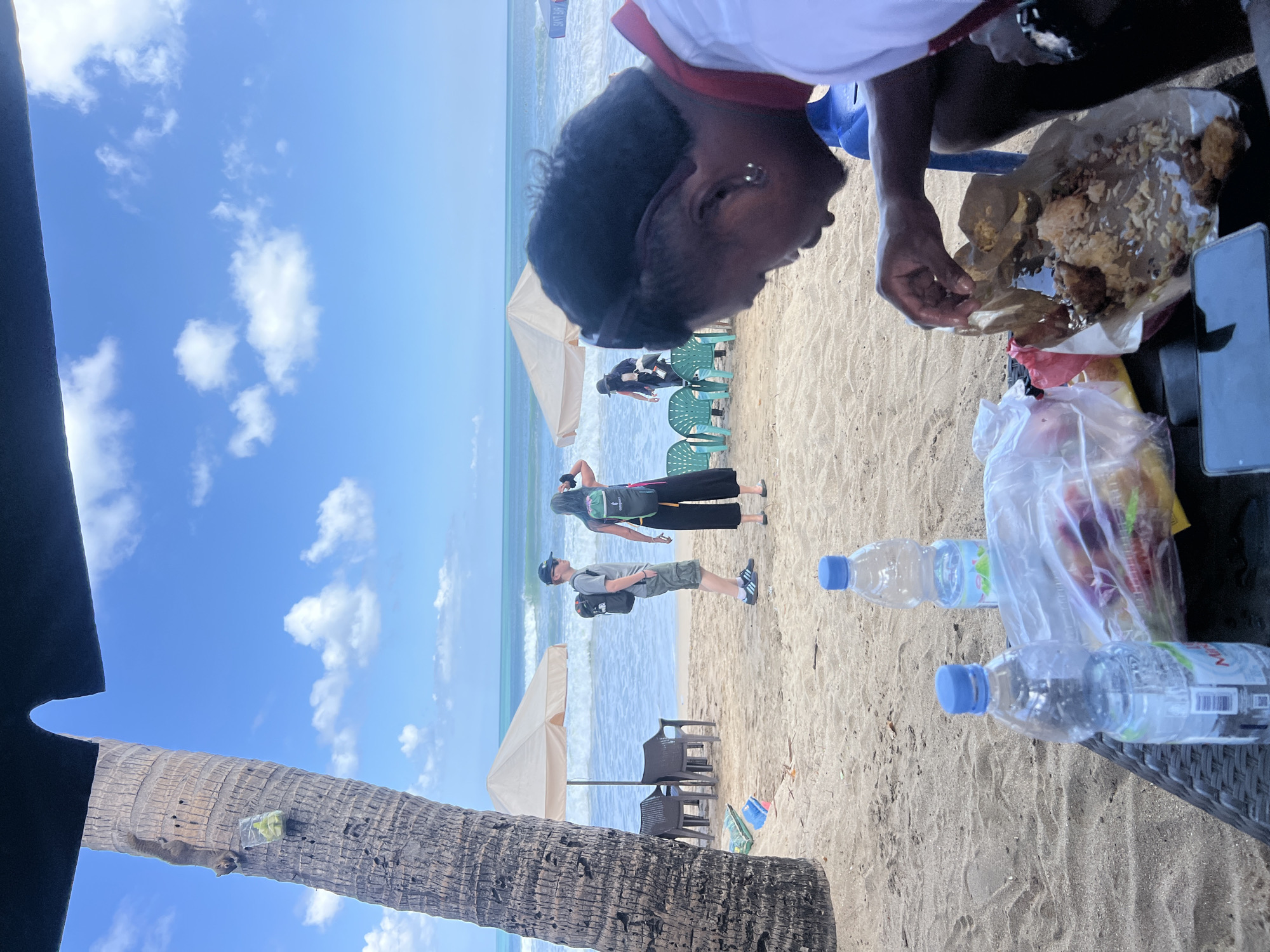 A group of people are enjoying a meal on a sandy beach in Kuta, Indonesia. The ocean waves are visible in the background, and there are several umbrellas and chairs set up on the sand. A person in the foreground is eating from a paper plate, while others stand and chat. A palm tree stands to the left of the frame, with a squirrel climbing its trunk. Two bottles of water and a bag of food are on a table in the foreground. The sky is blue with scattered clouds.