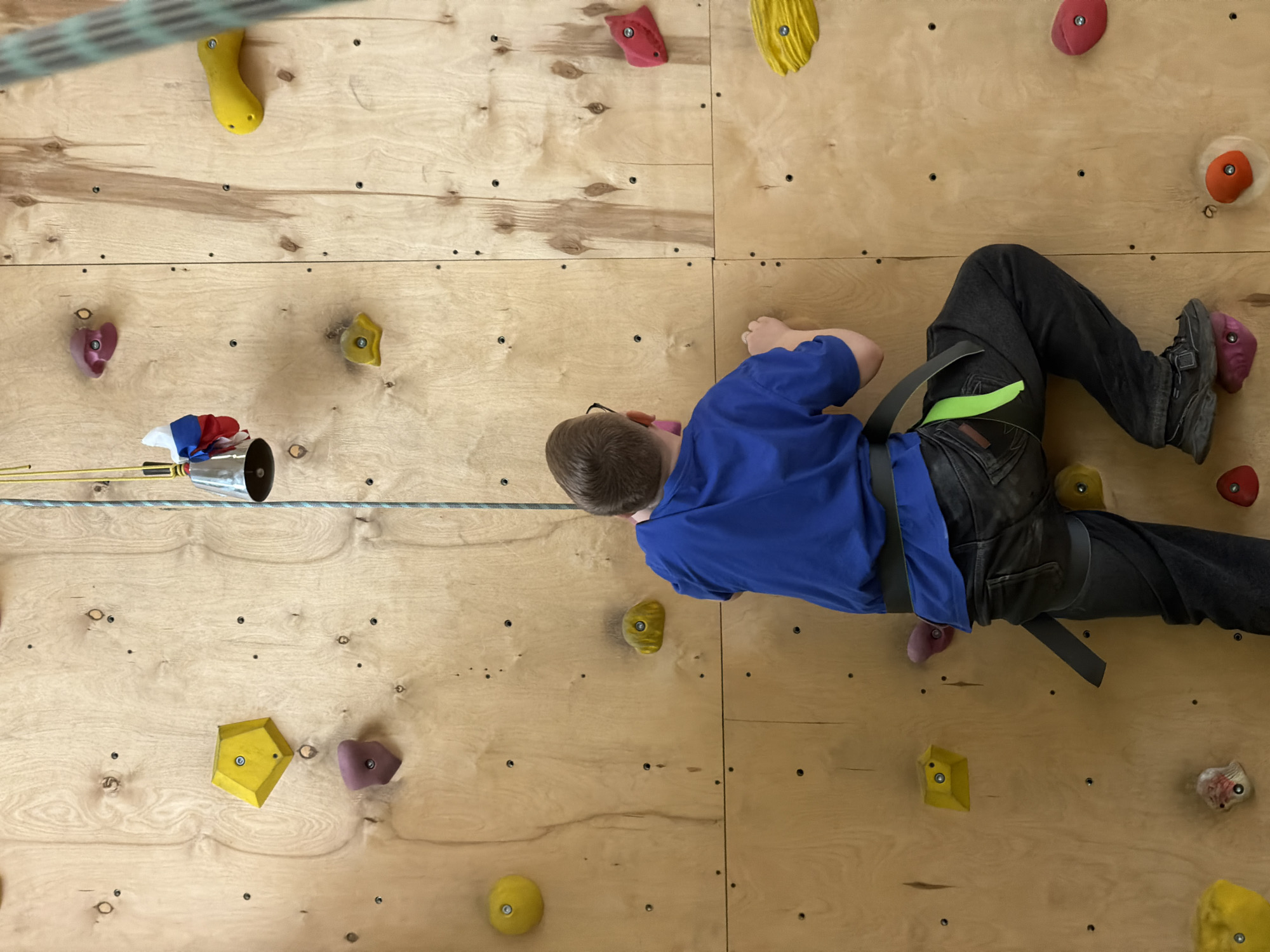 A boy in a blue shirt, dark jeans, and climbing shoes is on a climbing wall. He is wearing a climbing harness and is attached to a rope. The climbing wall is made of wood and has various colored climbing holds. A rope with a small bell with a Russian flag attached is also visible on the wall. The location is Izhevsk, Russia.