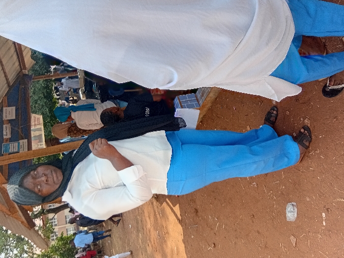 A woman in a hijab, white blouse, and blue pants is shown in a medium shot. She looks at the camera. A white cloth is draped above her, partially obscuring other seated figures. Other people sit behind her. The setting appears to be outdoors, with a dirt ground and a building in the background. The photo seems to be taken at a slight angle.