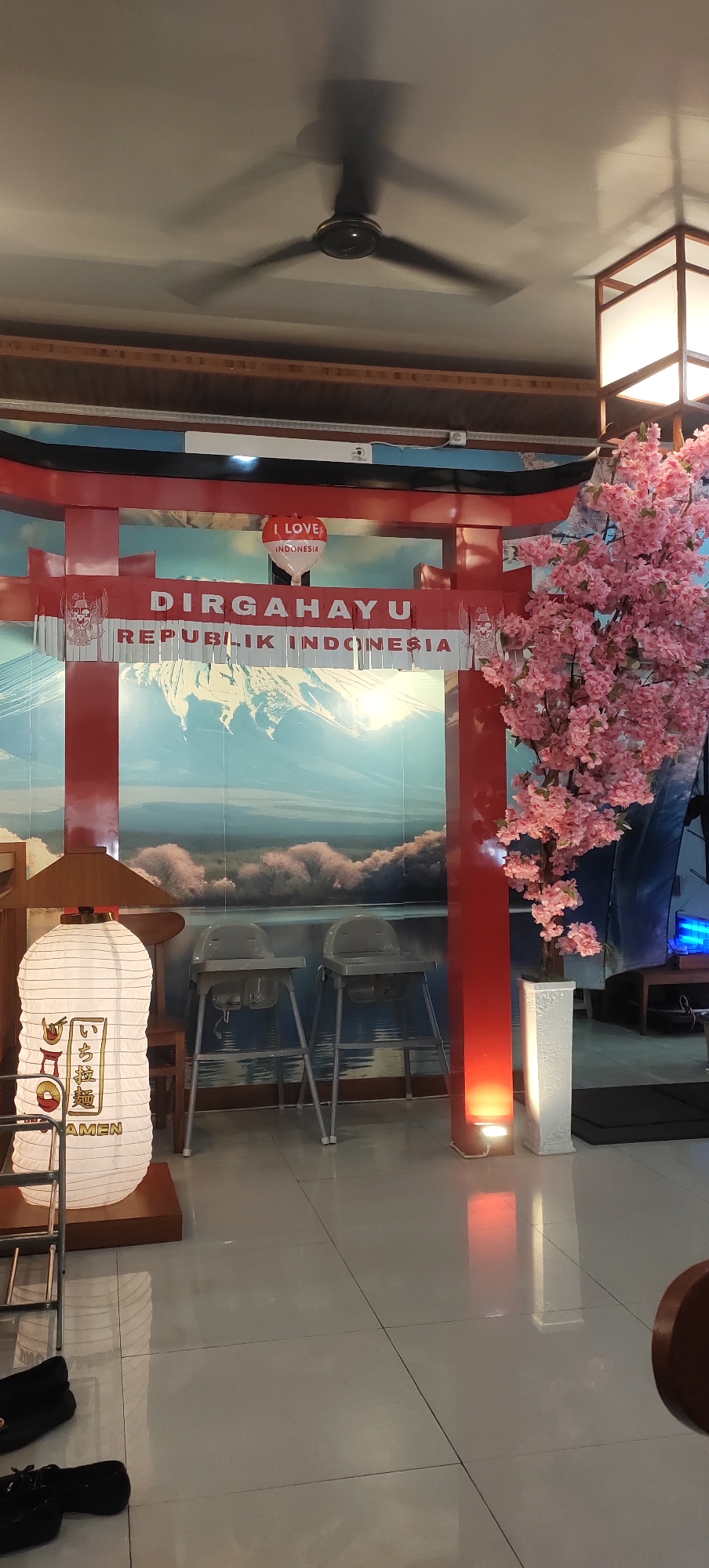 The image shows a festive display, likely for a national holiday, set up indoors. A large red torii gate, a traditional Japanese gate, serves as a backdrop with a painted mural of Mount Fuji behind it. The words "DIRGAHAYU REPUBLIK INDONESIA" are prominently displayed across the gate, indicating a celebration of Indonesian Independence Day. A heart-shaped balloon with "I LOVE INDONESIA" written on it is attached to the gate. To the right of the gate, a large artificial cherry blossom tree with pink flowers adds to the celebratory atmosphere. In front of the gate, a tall, white paper lantern with Japanese characters and the word "RAMEN" is positioned to the left. Two empty high chairs for children are placed in the center. The floor is tiled, and a ceiling fan is visible at the top of the image. The overall scene suggests a restaurant or a public space decorated for a national event. The provided crop hints do not offer specific details about the city of Kudus, Indonesia.
