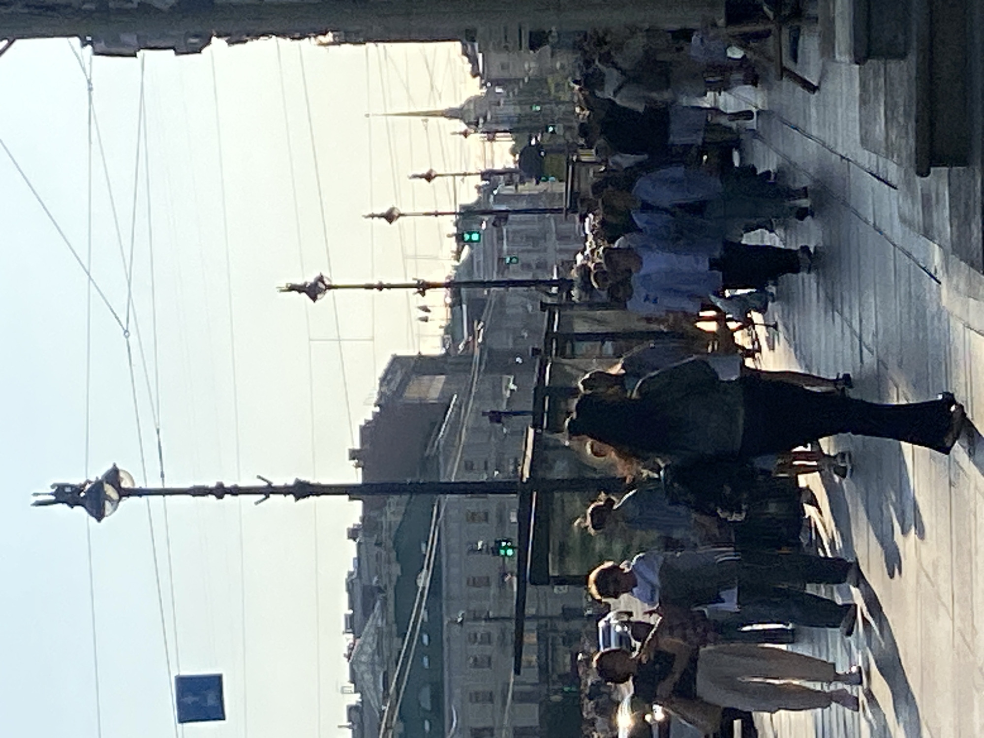 The image depicts a sunny day on a busy street in Saint Petersburg, Russia. Pedestrians walk along a wide sidewalk lined with buildings. The architecture suggests a historic city center. Several street lamps and traffic signals are visible. The lighting indicates late afternoon or early morning sun, casting long shadows on the pavement. A group of people is walking towards the camera, while others are seen further down the street. Some individuals are carrying bags, and one person is wearing a backpack. The overall scene is one of everyday urban life.