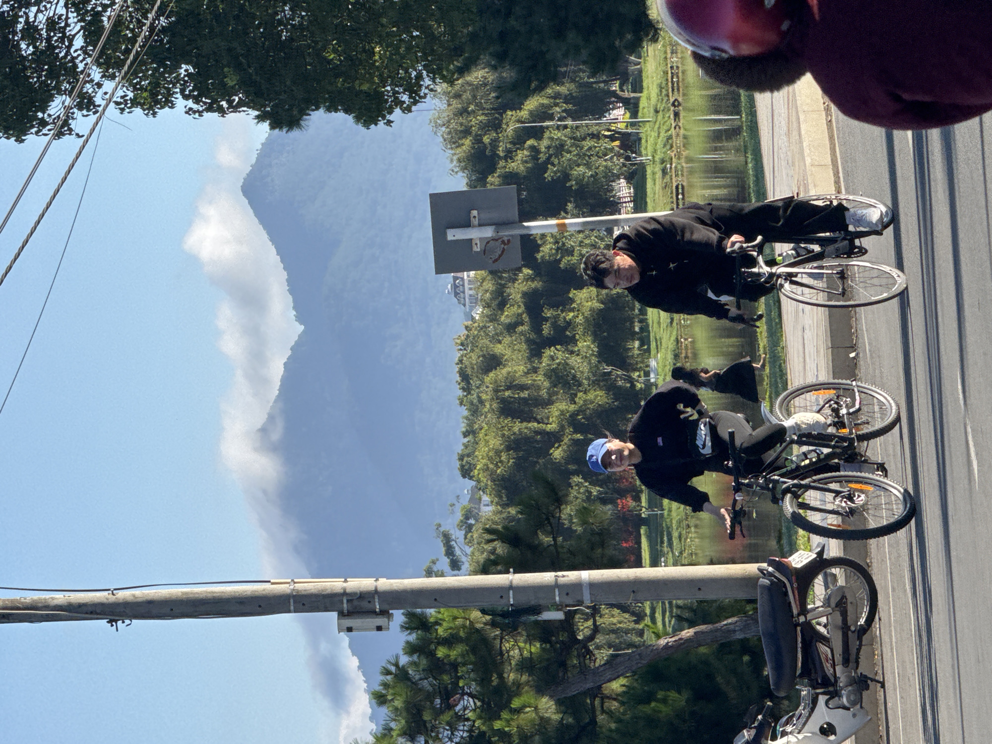 The image captures a scene in Da Lat, Vietnam, featuring two individuals cycling on a paved road. In the foreground, on the right, a person wearing a maroon helmet and fur-lined earmuffs is partially visible, facing away from the camera. To their left, a person in a black hoodie and pants, wearing white sneakers, is riding a road bicycle. Their hands are on the handlebars, and they are looking forward. Further to the left, another person wearing a blue baseball cap, black hoodie, and pants, also on a bicycle, is visible. This cyclist is also looking forward, with one hand on the handlebar. To the left of the cyclists, a motorcycle is parked. In the background, there is a body of water, possibly a lake or pond, with lush green trees lining its edge. Beyond the trees, mist-shrouded mountains form the backdrop, with a clear blue sky above. A utility pole stands prominently on the left side of the image, with wires strung across. A signpost is also visible behind the cyclist on the right. The overall ambiance suggests a bright, possibly morning or late afternoon, day.