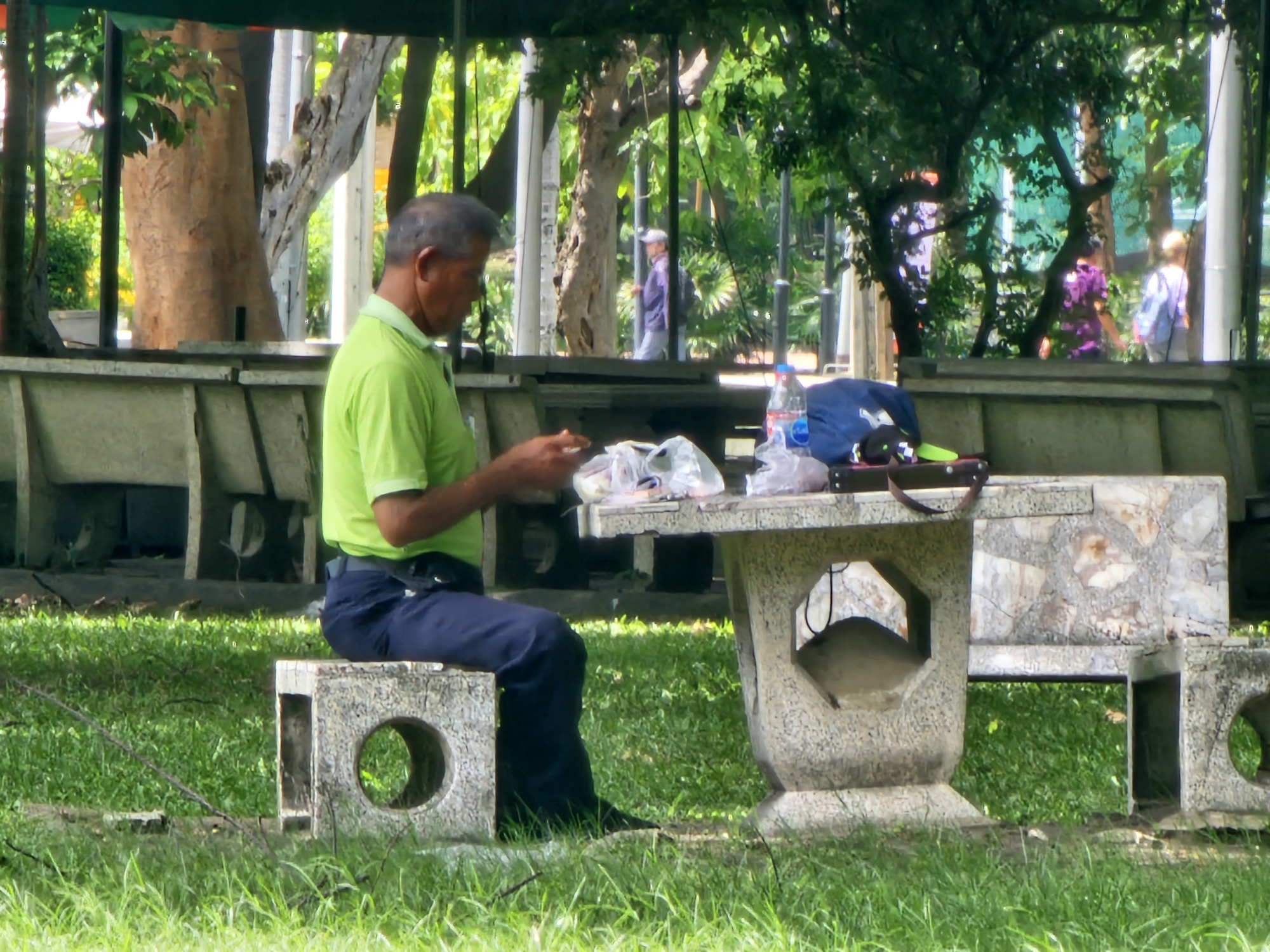 The image depicts a man in a green polo shirt and dark blue pants sitting on a concrete bench with a circular cutout in the front. He is holding some white plastic bags and appears to be sorting through them. In front of him is a stone table where more plastic bags and a blue bag are placed. To the right of the table, a water bottle is visible. The scene is set in a park-like environment with lush green grass and trees in the background. In the background, several people are walking, including one person in a purple shirt and another in a light-colored shirt with a backpack. The architecture of the benches and table suggests a public space in Bangkok, Thailand, likely a park or garden. The overall atmosphere is casual and relaxed, with the man engaged in a solitary activity.