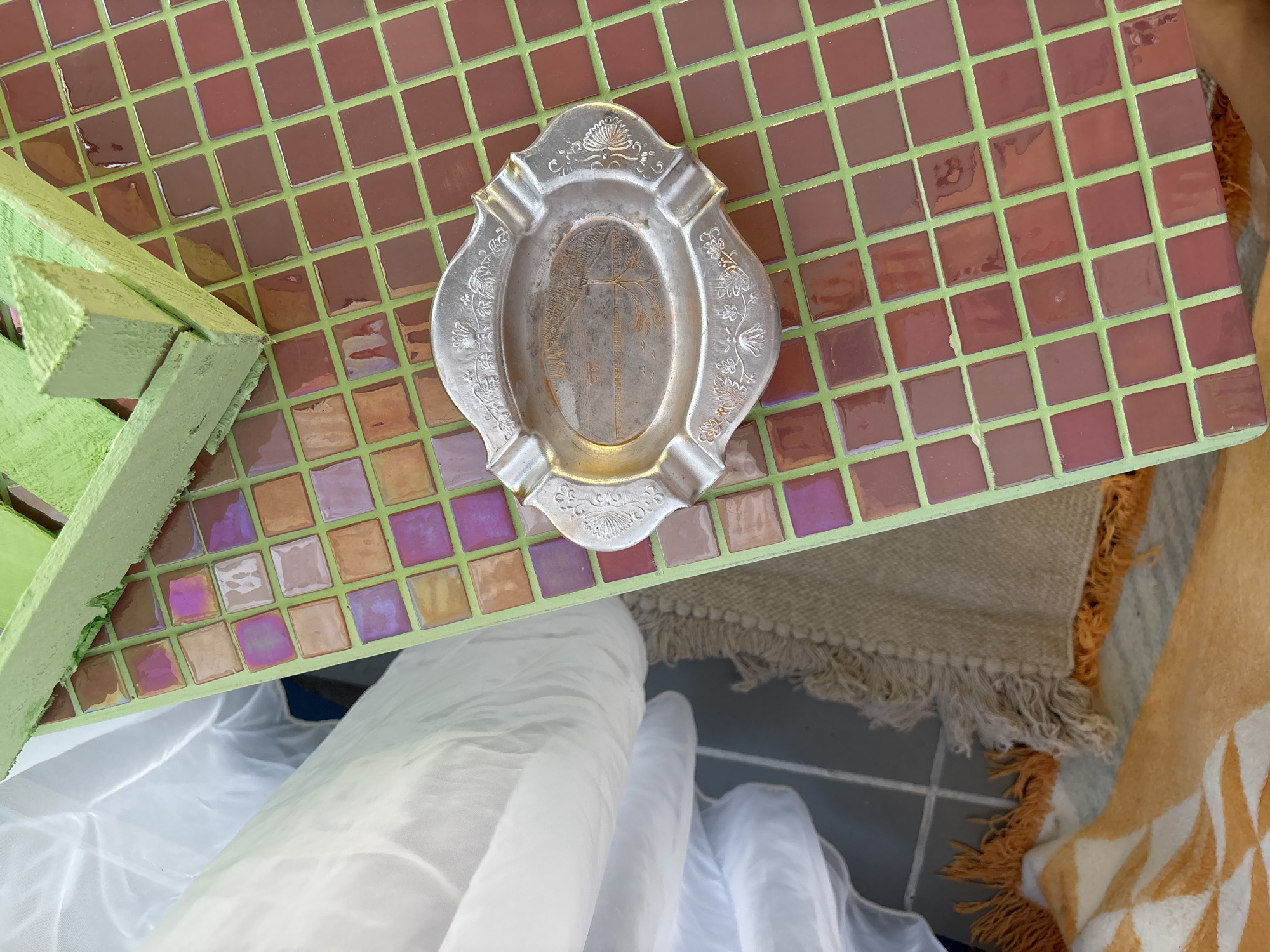 The image presents an overhead, slightly angled view of various objects on and around a tiled surface.
The primary subject is an ornate, silver-toned metal dish, likely an ashtray, centrally placed on a tiled tabletop. The dish has a decorative, scalloped edge with four indentations for cigarettes. Its surface is engraved with intricate floral and shell motifs, and its oval center features a distinct image resembling a stylized palm tree.
The dish rests on a mosaic-tiled surface composed of small, square tiles. The majority of these tiles are a glossy reddish-brown or maroon, set apart by bright light green grout lines. Towards the bottom left, some tiles display an iridescent sheen, reflecting shades of pink, purple, and orange.
To the left, a portion of a light green, roughly painted wooden structure, possibly a table leg or support, is visible. Below the tiled surface, a voluminous white, semi-transparent fabric is gathered. Further right and beneath, parts of two additional fabrics are visible: a light-colored, woven textile with thick fringes, and an orange and white patterned fabric, also with fringes, which could be a blanket or towel. A dark grey tiled floor with lighter grout lines is discernible beneath these items.
No people are present, and no specific activity is occurring. The lighting suggests it is daytime. There is no visible text within the image. The location context provided is Krasnoyarsk, Russia, though no explicit visual cues within the image directly indicate this geographical location.