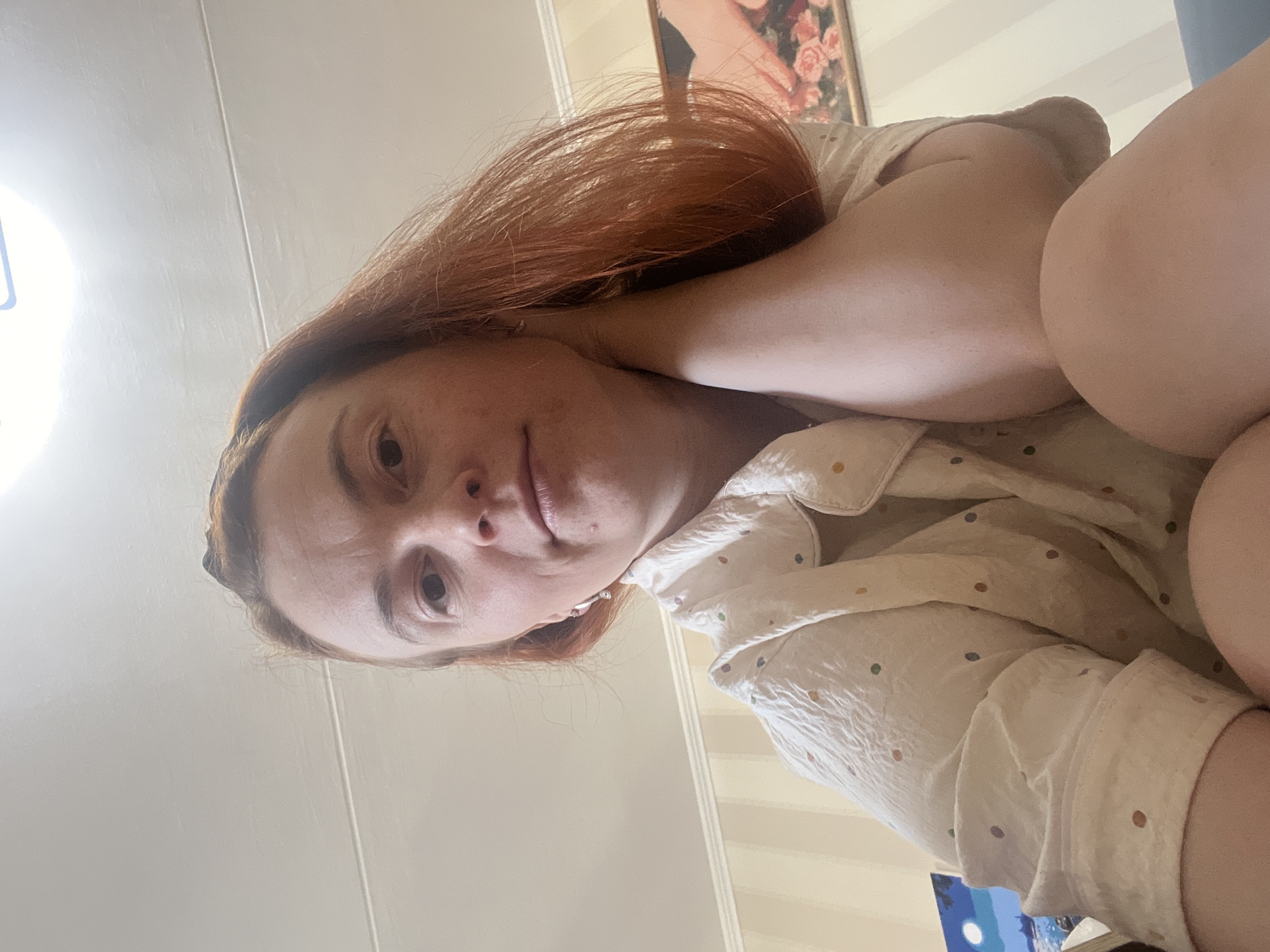 A selfie of a fair-skinned woman with reddish-brown hair. She is wearing a white shirt with colored polka dots and is laying down. Her left arm is bent and placed behind her head. In the background, there is a striped wall and a framed picture hanging on the wall. A portion of what appears to be a canvas with an image of a moon is visible at the bottom. The location is undetermined, and there is no direct indication of Chaikovsky, Russia.