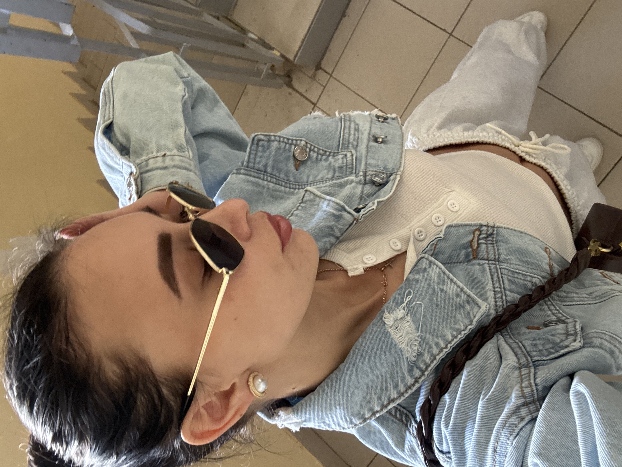 A woman is posing in a selfie with the camera positioned above her, looking up. She is wearing sunglasses with gold frames, a light blue denim jacket, and a white button-up top. She also appears to be wearing light grey sweatpants. The background is a tiled floor and a metal staircase railing. The location is vaguely identifiable as Chaikovsky, Russia.