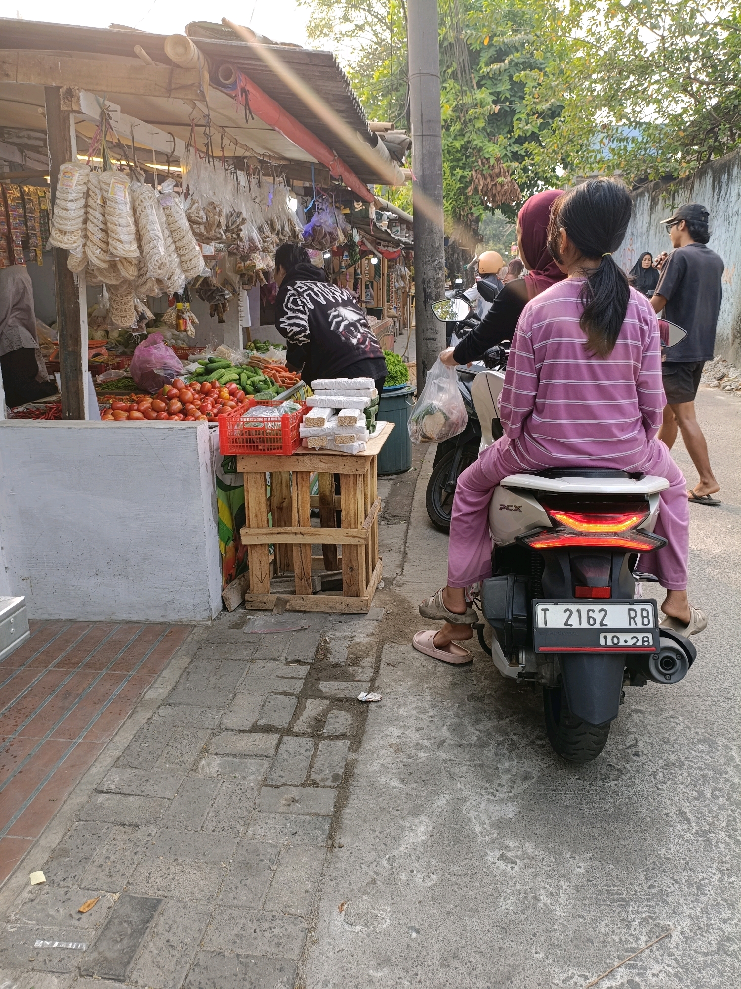 In Tangerang, Indonesia, a roadside market bustles with activity. A woman in a purple striped outfit rides as a passenger on a scooter, her legs dangling on either side of the vehicle. She faces away from the camera, her hair pulled back in a ponytail. Next to her, another person in a red headscarf also faces away. A third person, wearing a black hoodie, appears to be interacting with the market stall. The stall itself is laden with goods, including an abundance of tomatoes and cucumbers, as well as bagged snacks and other items. The ground is paved with concrete and brick, showing signs of wear and tear. A wooden stand holds a red crate filled with what appears to be food items wrapped in white packaging. The scene is bathed in the warm glow of sunlight, hinting at either early morning or late afternoon.