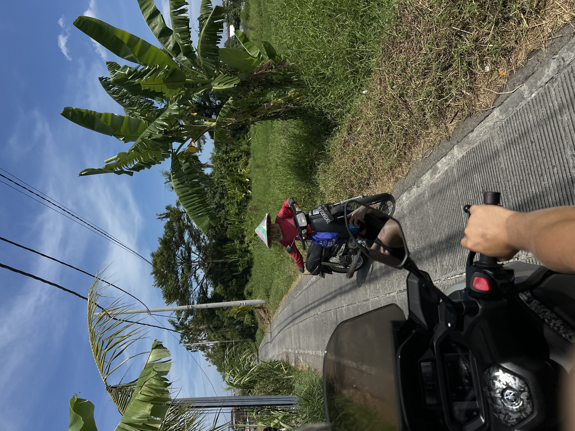 A view from a moving motorcycle on a concrete road in North Kuta, Indonesia, with a person wearing a traditional Asian conical hat and a red long-sleeved shirt riding a motorcycle in the same direction. The motorcycle has cargo bags strapped to the back, and a license plate visible with letters "DK6555FB". Lush green foliage, including banana trees and tall grasses, lines the road. The sky is blue with scattered clouds, and overhead power lines run parallel to the road. The foreground shows the handlebars and part of the dashboard of the motorcycle from which the picture is taken, along with a hand on the grip.