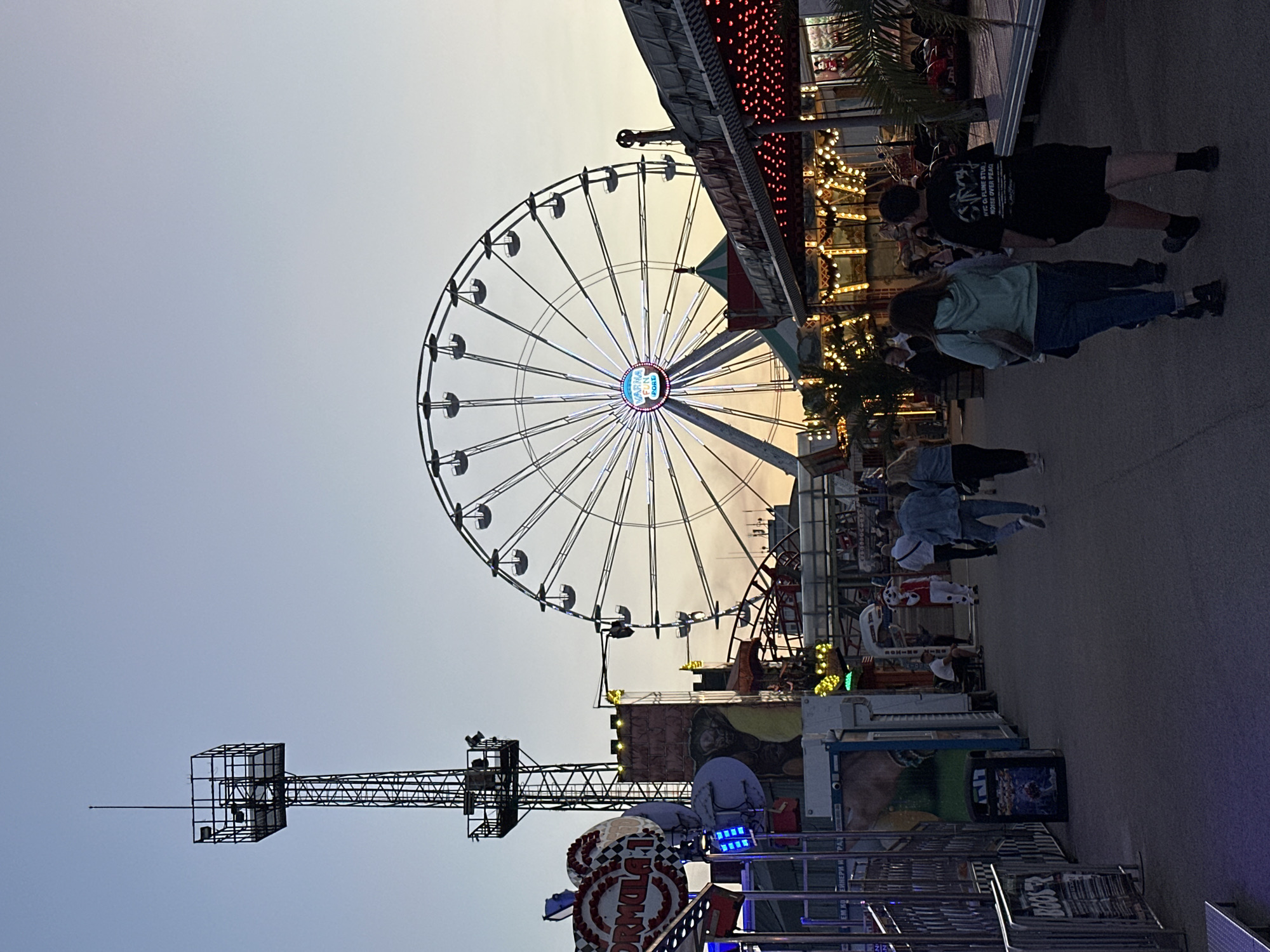 The image depicts an amusement park in Varna, Bulgaria. A large Ferris wheel dominates the background. A drop tower ride is visible in the lower left. Several people are walking along a pathway near various game booths. The sky suggests either dusk or dawn.
