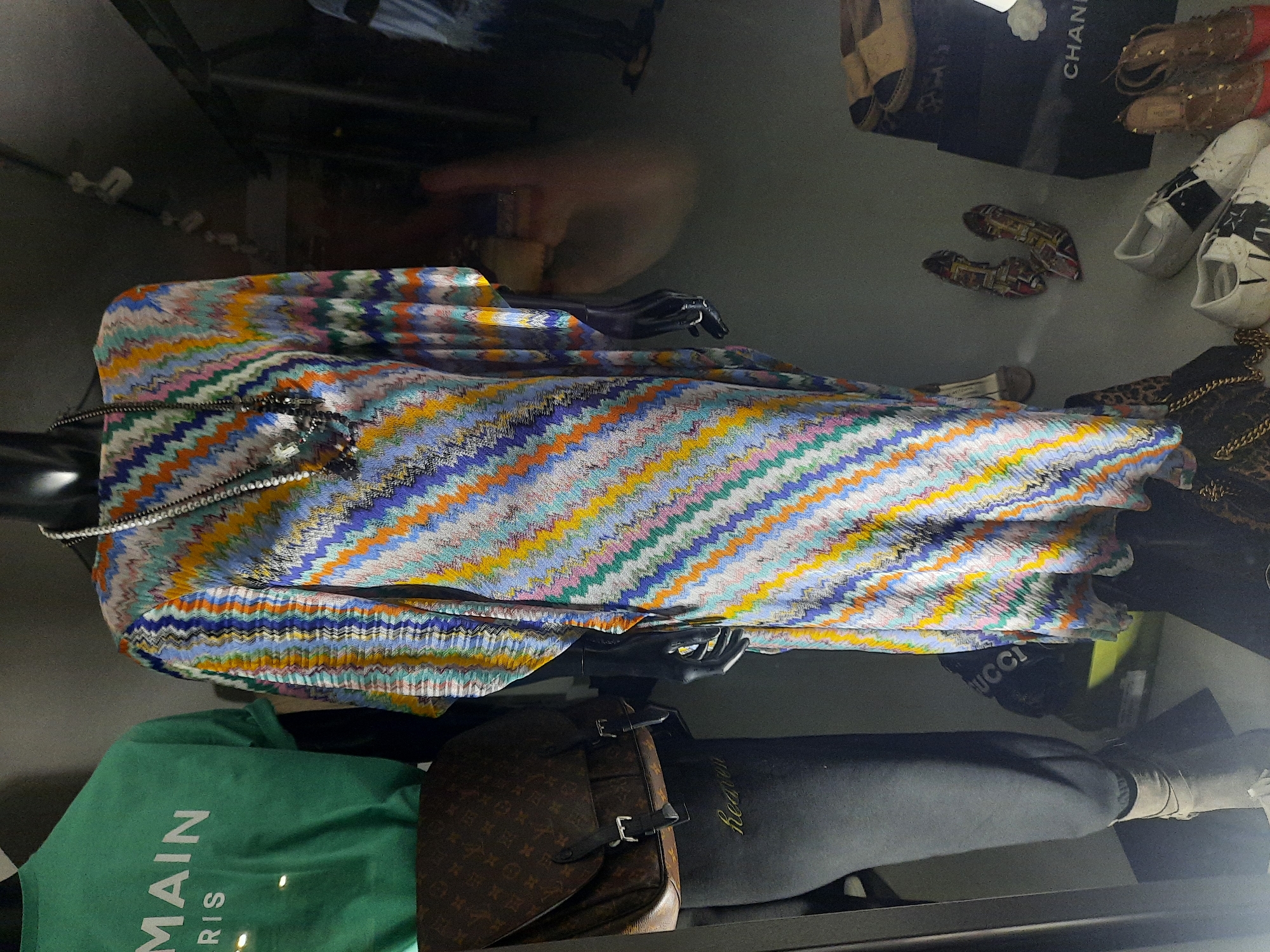 The image shows a display in a retail environment, likely a shop window or an in-store display, in Rosenheim, Germany. A mannequin on the left is wearing a green t-shirt with white text that reads "MAIN PARIS". A brown Louis Vuitton messenger bag is slung across its shoulder. Below the bag, dark gray sweatpants with the word "heaven" embroidered in gold script are visible. To the right, another mannequin is dressed in a colorful, zigzag-patterned kaftan or maxi dress. It is adorned with a long, beaded necklace. In the background, various shoes and shopping bags are displayed, including a black Chanel box. The lighting suggests an indoor setting, possibly a boutique or department store. The overall scene is a curated retail presentation of clothing and accessories.