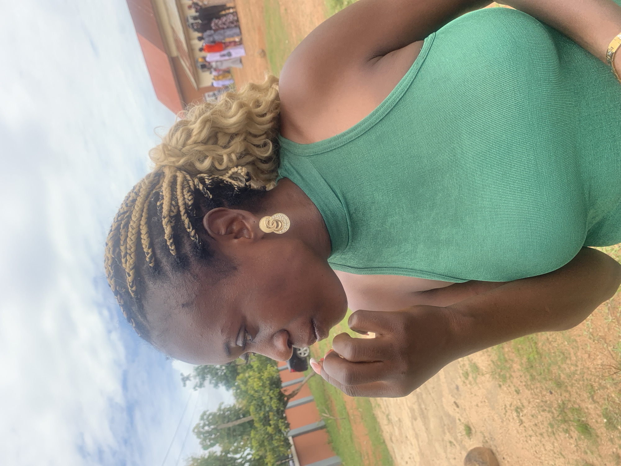 The image captures a woman in a green tank top, with blonde-streaked braids and large, circular gold earrings, looking to her left and touching her mouth with her fingers. She appears to be outdoors, with a dirt and grass ground beneath her and a cloudy sky above. In the background, several people in traditional attire are gathered in front of a building with a red roof, suggesting a possible event or ceremony. The scene is likely set in Duk, Nigeria, given the context provided.