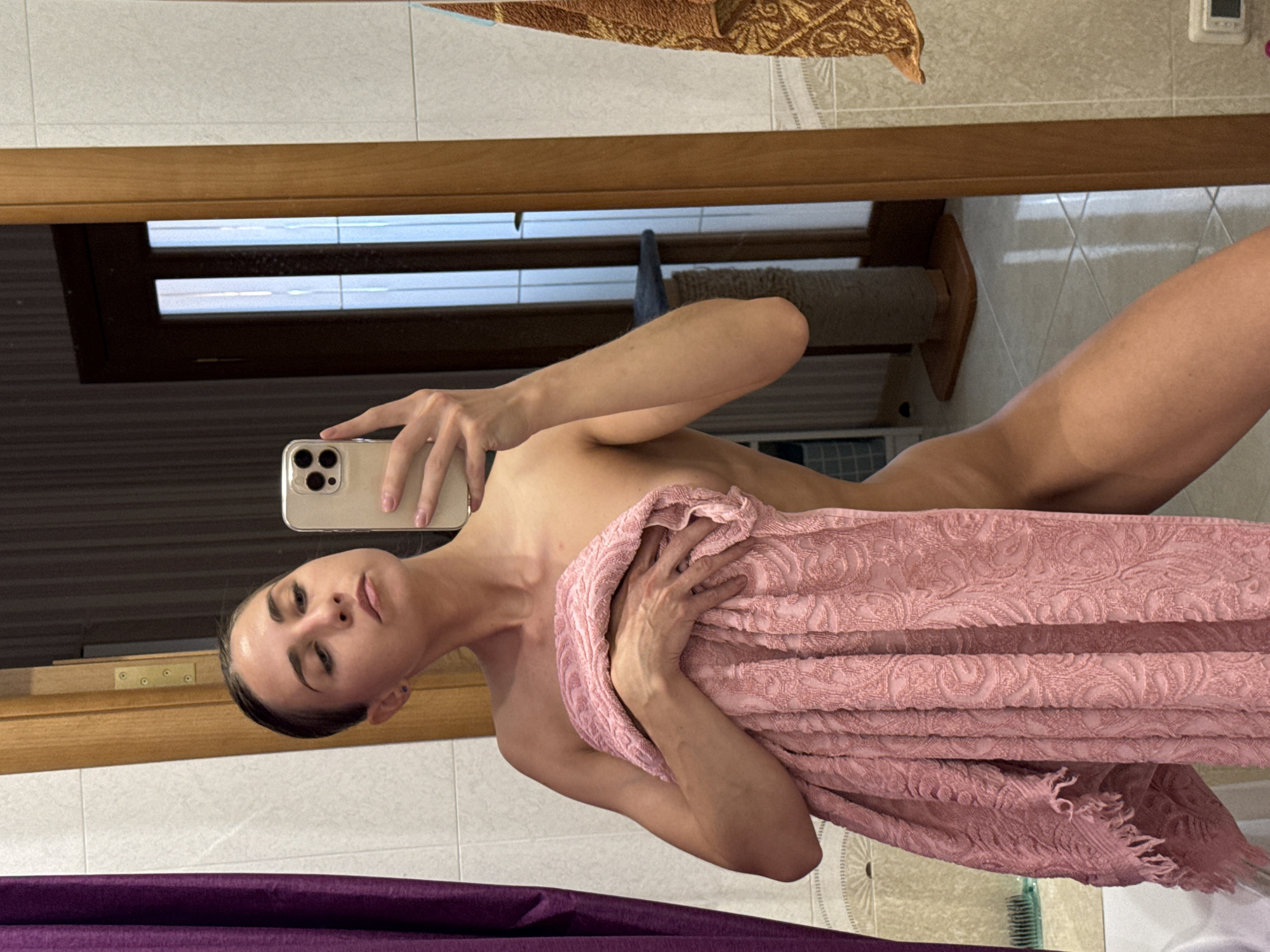 A young woman is captured in a mirror selfie, with the image rotated 90 degrees clockwise. She holds a light-colored smartphone with multiple cameras in her left hand, angling it to photograph her reflection. Her dark hair is pulled back, and she looks towards the camera with a slight pout and prominent eyebrows. She is draped in a textured, light pink towel, which she holds with her right hand to cover her chest. Her bare shoulders and arms are visible.
The reflection in the mirror reveals an indoor setting, likely a bathroom or dressing area. A light brown wooden frame, possibly a door or window, runs horizontally across the upper half of the image. Through the reflection of what appears to be a window, horizontal blinds or slats are visible, suggesting natural light from outside. In the upper right quadrant of the reflection, a beige cat scratching post stands, and above it, a white wall-mounted thermostat or control unit is visible. A decorative brown and gold patterned fabric hangs in the background. The floor is tiled, and a green comb or brush is partially visible at the bottom of the frame, near a dark purple or maroon fabric element, possibly a towel or bathmat. No visible text is present in the image.