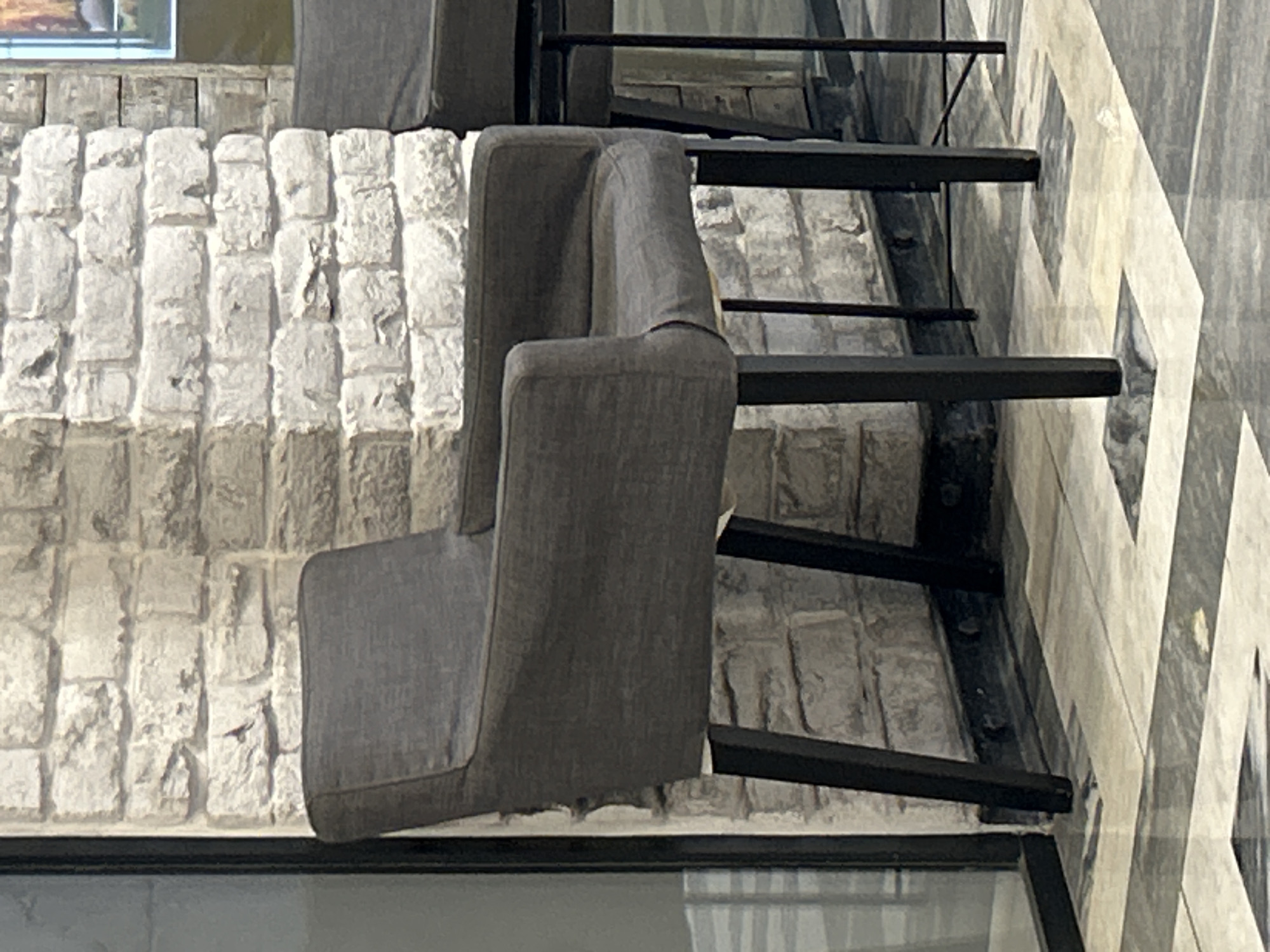 The image displays an interior setting in the city of Unknown, Unknown. A dark gray upholstered armchair with black, tapered legs is centrally positioned, angled slightly towards the left. The chair features a high back and integrated armrests. It stands on a polished floor composed of large, reflective tiles arranged in a pattern of alternating light and dark squares. Behind the chair, a wall constructed from irregularly shaped, light-toned stone or brick blocks is visible, exhibiting distinct mortar lines and textured surfaces. A small, dark, linear mark is present on the wall above the chair's backrest. To the left of the chair, a black-framed glass panel, possibly part of a partition or door, reflects ambient light. Along the base of the stone wall, a dark, horizontal element, likely a baseboard or railing, is visible with several circular fixtures. A second, identical dark gray chair is partially visible on the right, with only its back and some legs in frame. No individuals are present in the scene.