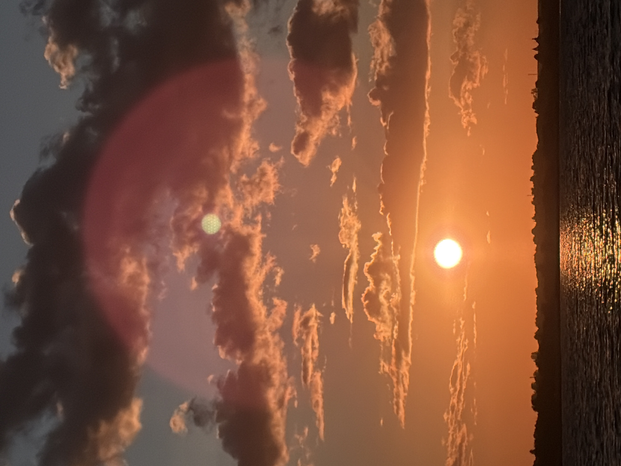 The image depicts a sunset over a body of water, likely a river or lake, in Vidogoshchi, Russia. The sky is filled with dramatic clouds tinged with orange and pink hues from the setting sun. Two suns are visible in the sky, one in the lower half of the frame, appearing as a bright, distinct orb, and another smaller, fainter orb higher up. A large, soft, pinkish-red halo effect is present in the upper portion of the sky, obscuring some of the clouds. Silhouetted against the vibrant sky is a distant tree line or cityscape. The water in the foreground reflects the golden light of the sun.