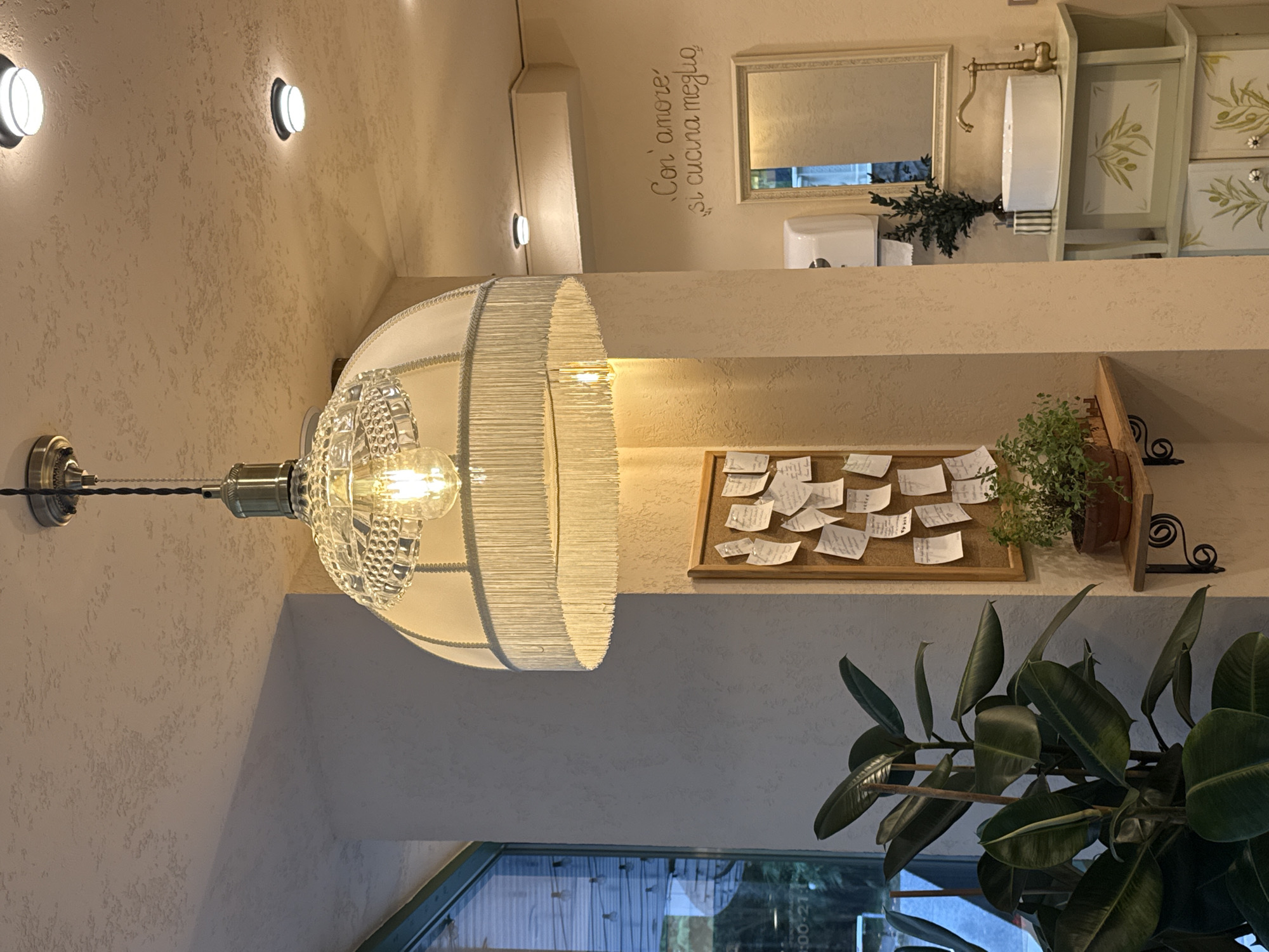 The image depicts the interior of a brightly lit establishment, possibly a cafe or restaurant, with a focus on decorative elements and a sense of a well-maintained space. Prominently featured is a vintage-style pendant lamp with a glass shade and fringe, hanging from a textured ceiling. Two circular ceiling lights are also visible. To the left, a large rubber plant with dark green leaves adds a natural element. A corkboard covered in handwritten notes is mounted on a wall. A small potted plant sits on a wooden shelf supported by ornate brackets. In the background, a vanity with a white basin, a brass faucet, and decorative cabinets is visible, along with a mirror and a soap dispenser. The wall features the Italian phrase "Con amore si cucina meglio" (With love, one cooks better) written in cursive. The overall aesthetic suggests a cozy and inviting atmosphere. There are no individuals visible in the image. The location is identified as Sochi, Russia.