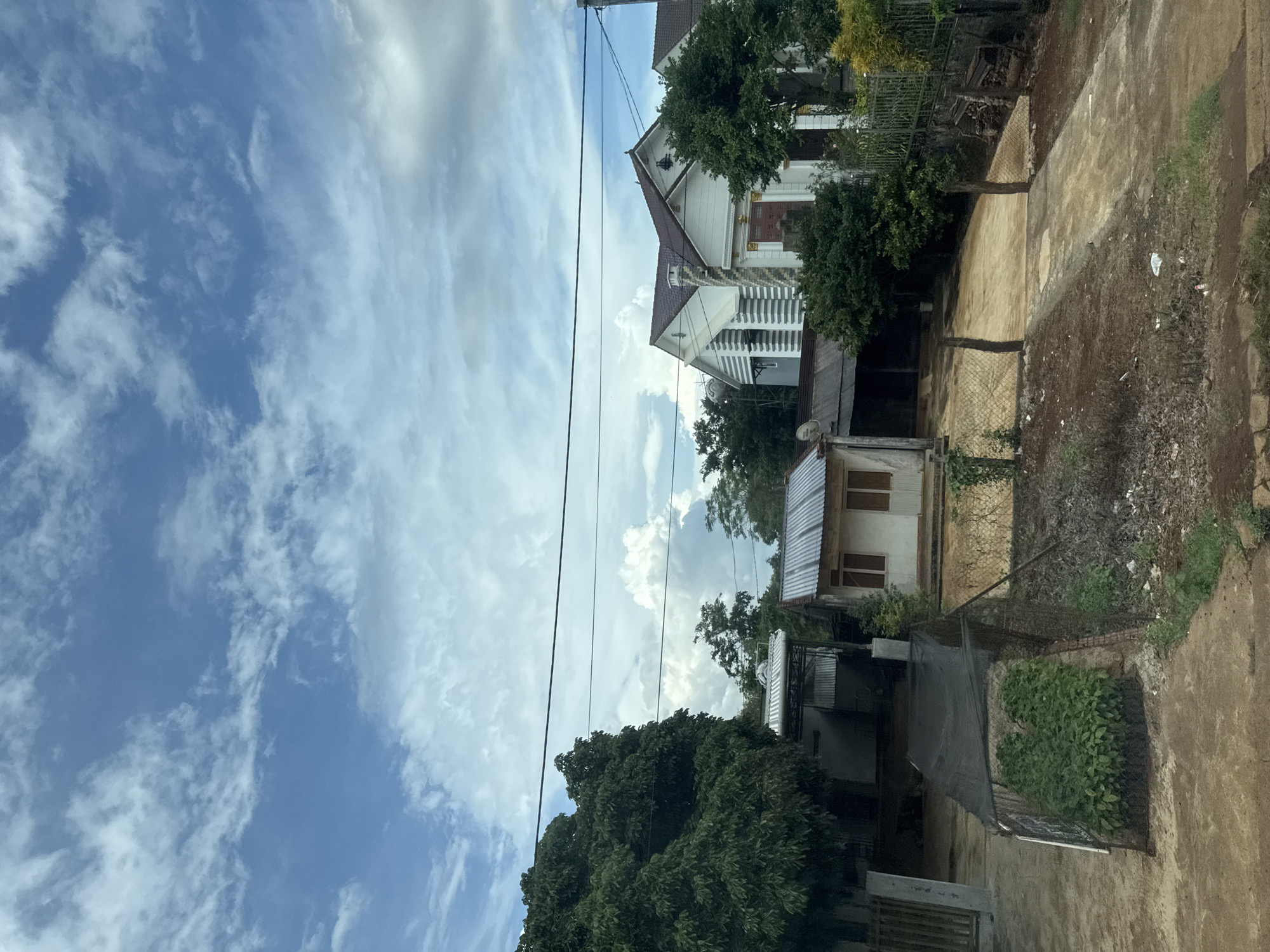 This is a landscape photograph of a rural scene in Chư Sê, Vietnam, taken during the daytime under a cloudy sky. The image is oriented vertically, with the sky occupying the left half and the ground and structures on the right.
The scene features several houses and buildings, some appearing more modern with white exteriors and decorative elements like stairs and railings, while others are simpler with corrugated metal roofs. Vegetation, including lush green trees and shrubs, is abundant throughout the scene. Power lines stretch across the frame, indicating the presence of infrastructure.
The ground is a mix of dirt, gravel, and concrete paths. A chain-link fence is visible, dividing parts of the property. Small piles of debris and some scattered litter are present in the foreground. The overall impression is of a lived-in, somewhat unkempt but functional residential area. The clouds suggest a possibly humid or overcast day. There is no visible text in the image.