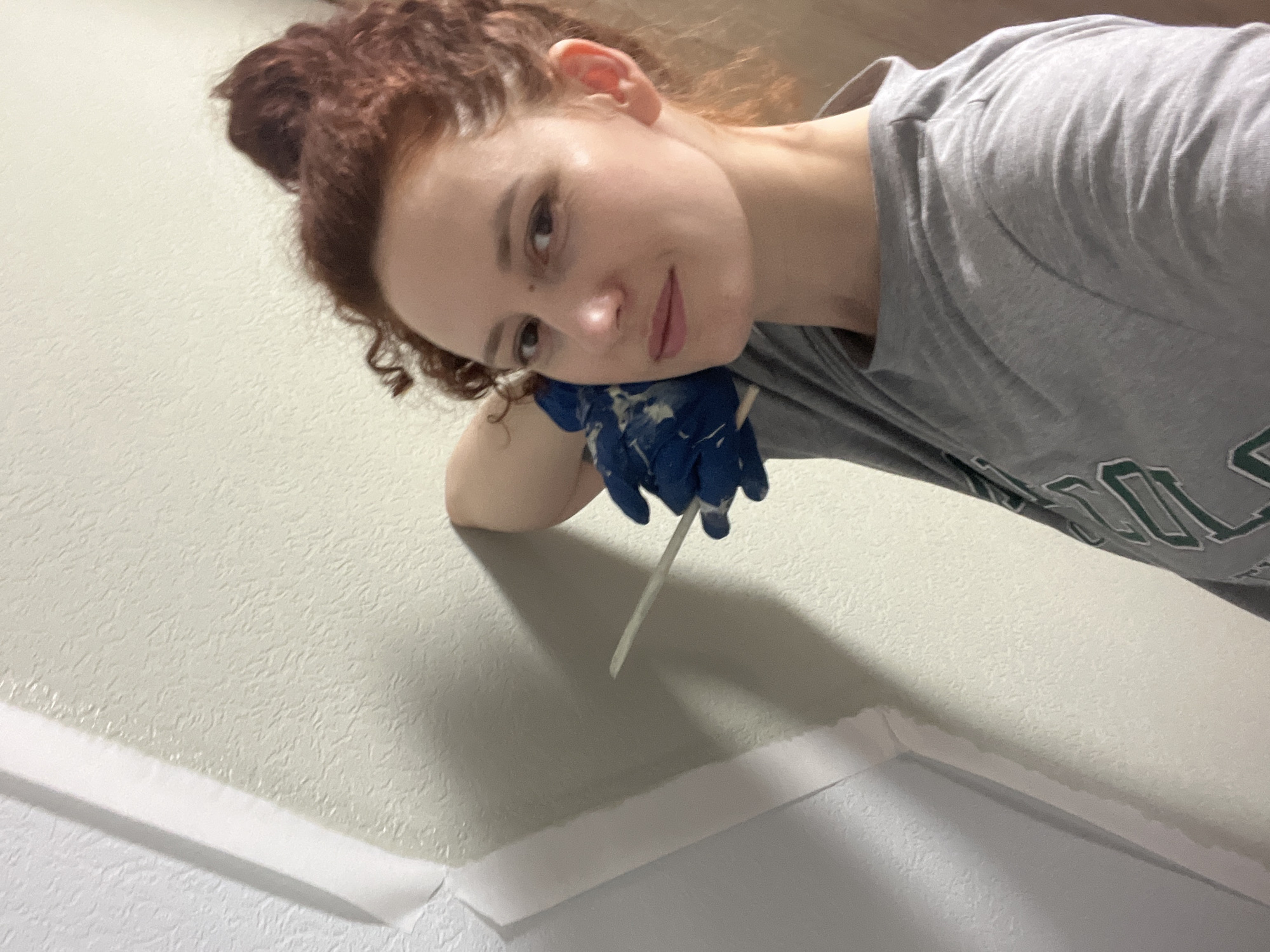 A selfie shows a woman with red curly hair in a high bun, wearing a gray t-shirt and blue rubber gloves, applying white caulk along a wall. The wall is painted a textured light beige color and has white trim along the bottom edge. A narrow stick is held in her gloved hand, apparently used for smoothing the caulk. The background is blurred and the location is not discernable from the image. There is no indication of the city of Saint Petersburg, Russia in the image.