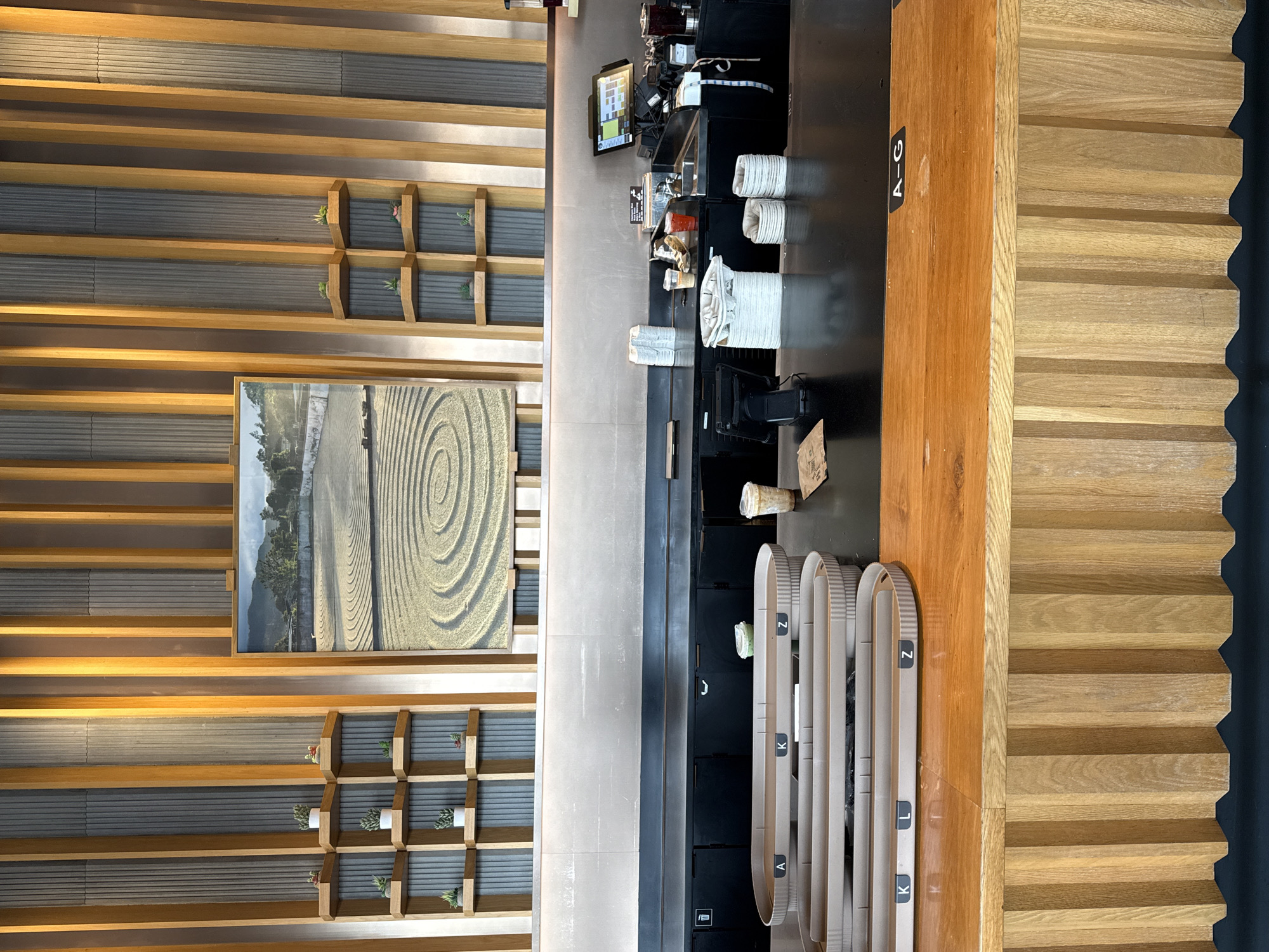 A vertically oriented indoor shot depicts the ordering area of a restaurant, possibly a cafe or coffee shop, likely located in Palm Springs, United States. The decor is dominated by a horizontal slat design in warm wood tones, which covers a large portion of the wall and extends to what appears to be a wall or partition. This slat design incorporates small square shelving units with minimalist plant decorations. A framed artwork featuring a spiral design is prominently displayed on the left-hand wall.
To the right, a dark countertop holds various service items such as stacks of cups, coffee-making equipment, and a point-of-sale system with a visible screen. The countertop transitions into a wooden barrier with signage reading "A-G." Below the countertop, there are several curved dividers labeled with letters, possibly indicating different order pick-up zones. The overall setting suggests a modern, well-lit commercial space.