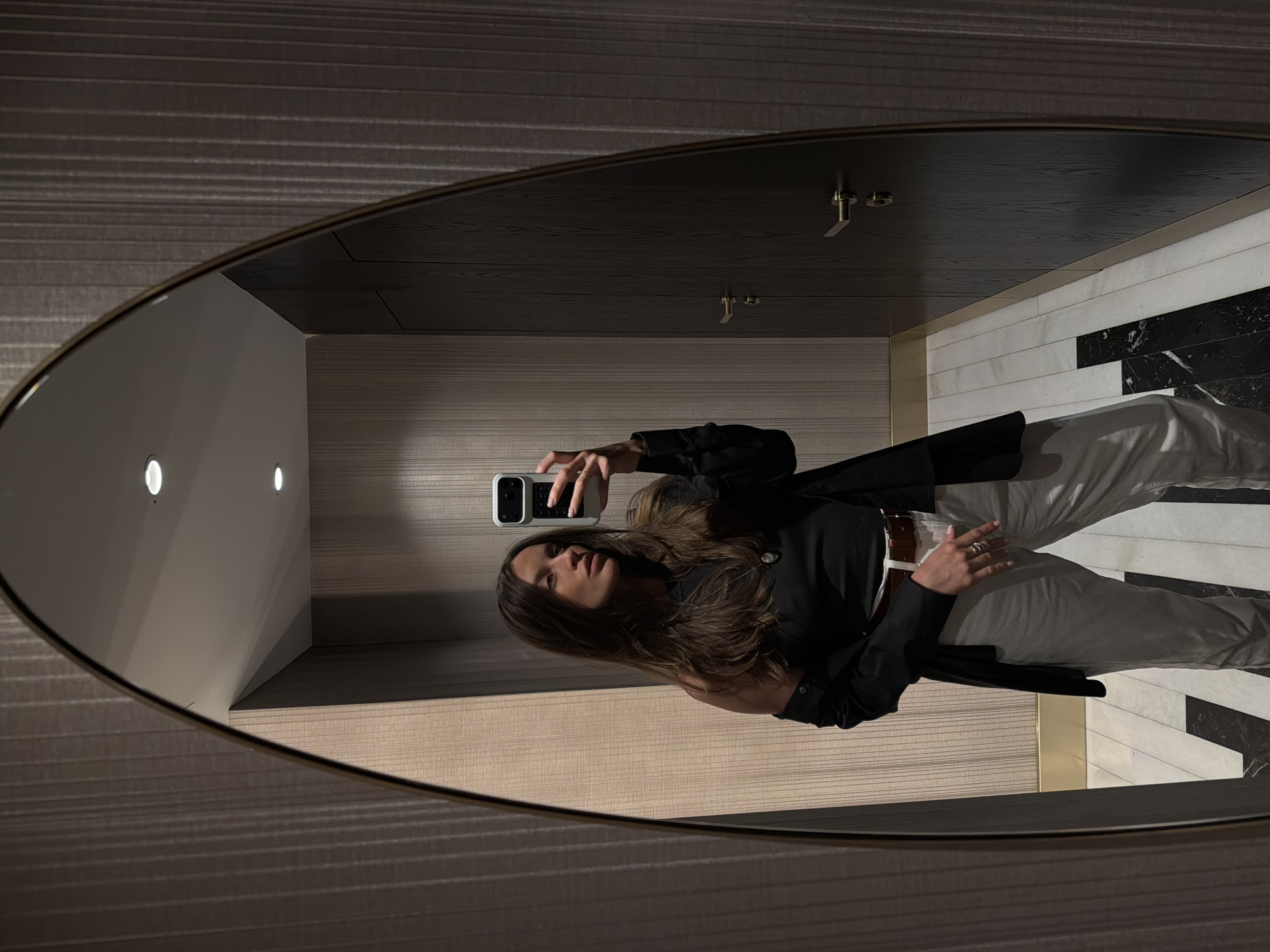 The image shows a person taking a selfie in a mirror. The mirror is oval-shaped with a metallic frame and reflects a room with a modern design. The walls are covered in a striped pattern, and the ceiling appears to be made of dark wood. The floor has a black and white marble design. The person in the reflection is wearing a black top and white pants and holding a white smartphone. The location appears to be a restroom or dressing room. It is impossible to identify the precise location in Budva, Montenegro, without additional context.