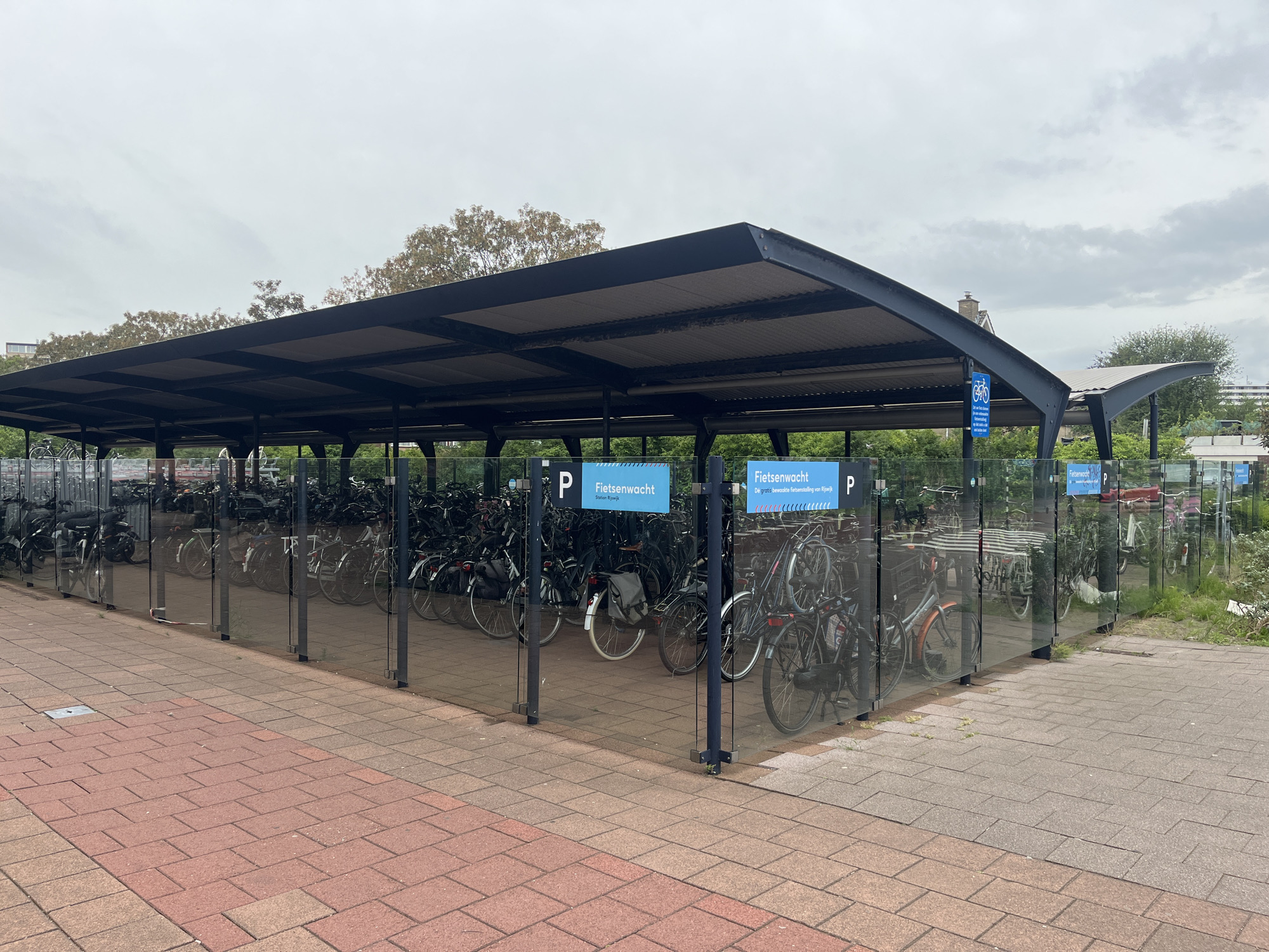 The image shows a covered bicycle parking facility with a modern design. The structure has a dark-colored, corrugated metal roof supported by black metal beams and pillars. The sides of the parking area are enclosed by clear glass panels, allowing a view of the bicycles parked inside. The bicycles are neatly arranged, with many of them appearing to be of the commuter or city bike style. Some of the bicycles have baskets or bags attached.
There are blue signs with white lettering visible on some of the pillars. One sign reads "Fietsenwacht" which translates to "Bicycle Watch" or "Bike Guard" in English, and also includes "Gratis onbewaakte fietsenstalling van Rijswijk", meaning "Free unsupervised bicycle parking of Rijswijk". Another sign shows a white bicycle symbol on a blue background.
The ground in front of the parking facility is paved with interlocking bricks in a reddish-brown color. To the right and behind the structure, there are green trees and foliage, suggesting an outdoor setting. The sky is overcast with gray clouds. The scene appears to be a public area, possibly near a train station or a busy urban location, given the presence of a large bicycle parking facility. There are no people visible in the image.