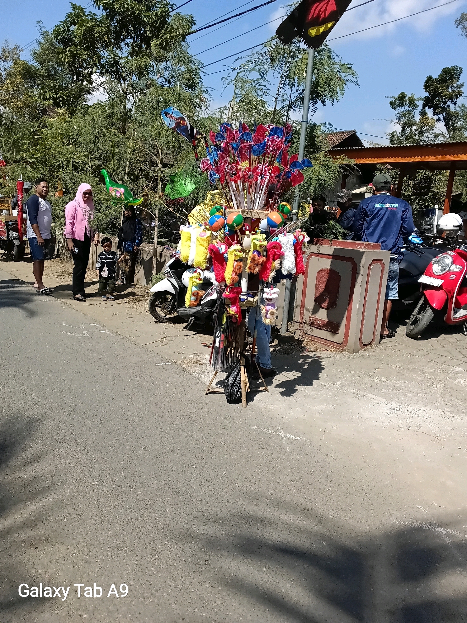 In the city of Bendo, Indonesia, a street vendor stands with a cart filled with colorful toys, including kites, pinwheels, and plush animals. A group of people are gathered around, some looking at the toys, while others are on motorcycles. A white scooter is parked to the left of the cart, and a red scooter is parked to the right. The street is paved with asphalt, and there are trees and buildings in the background. The sky is clear and blue, with a few clouds. The vendor appears to be interacting with a potential customer, a woman in a pink hijab.