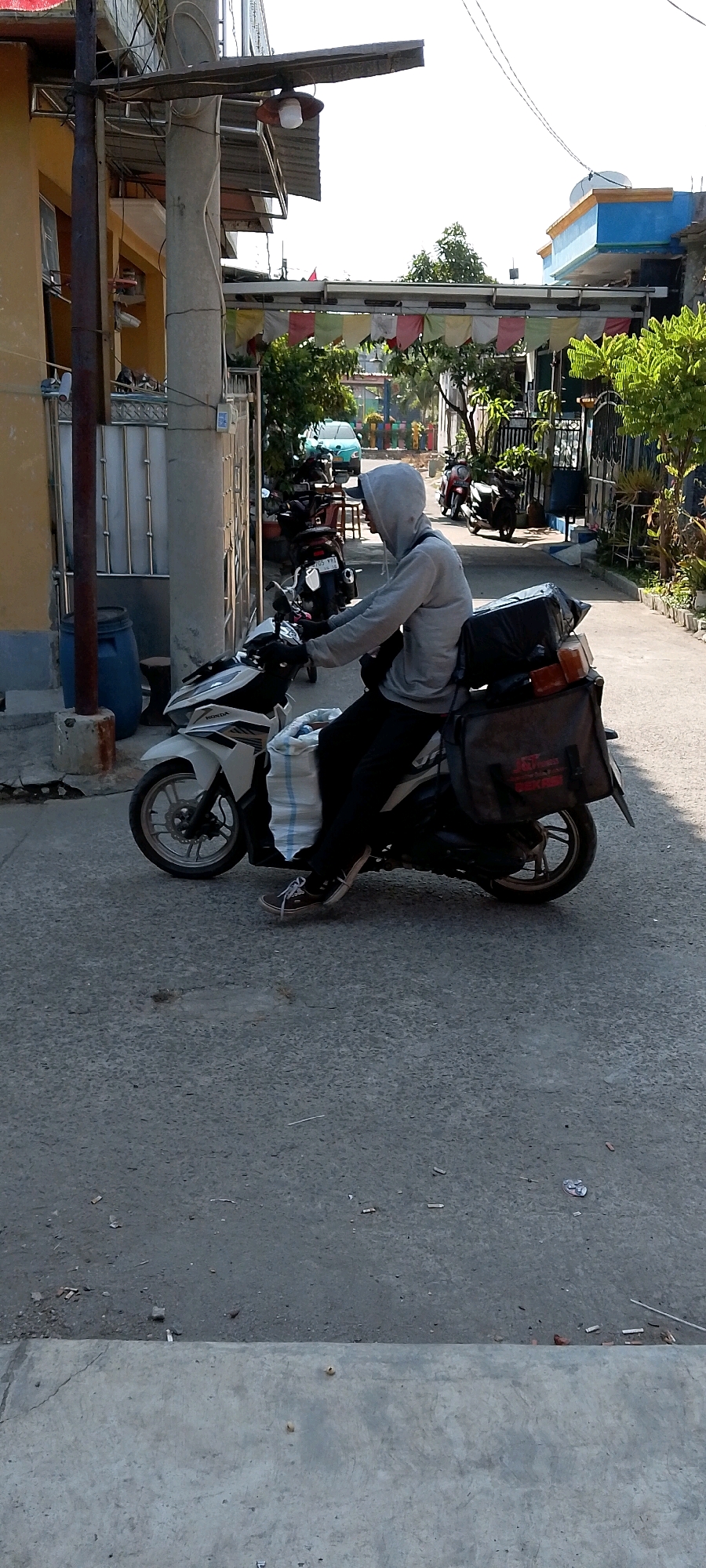 A person wearing a grey hooded sweatshirt and dark pants is riding a white and black scooter. The scooter is carrying a large white bag on the front and a black bag on the back. The person appears to be looking forward as they ride down a paved street. The street is lined with buildings and vegetation. In the background, there are other scooters and a light blue car. The scene appears to be in a residential area, likely in Satriamekar, Indonesia, given the architectural style and vegetation. The lighting suggests it is daytime, with sunlight casting shadows on the ground. There is a concrete structure on the left side of the image, possibly a utility pole with attached electrical wires and a light fixture. A blue plastic barrel is visible near the concrete structure. Colorful flags are strung across an overhead structure in the middle ground.