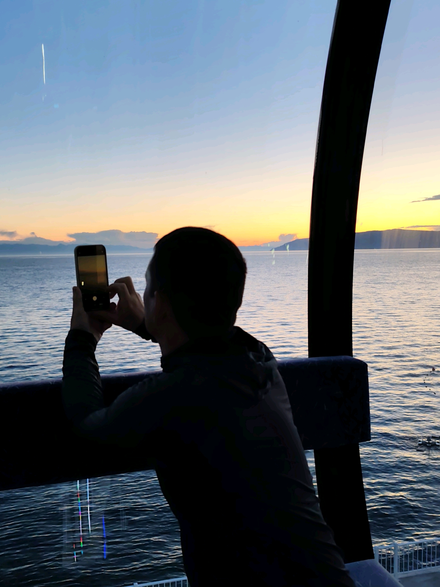 A person is silhouetted inside a gondola, holding a smartphone to capture a sunset over a body of water. The sky is a gradient of blue and orange, with distant mountains visible on the horizon. The water is calm, with ripples reflecting the sky's colors. The gondola has a dark, curved frame, and the person is wearing a dark jacket. The image appears to be taken in Listvyanka, Russia, as evidenced by the distinctive gondola likely belonging to the Baikal Cable Car and the surrounding landscape characteristic of the area.