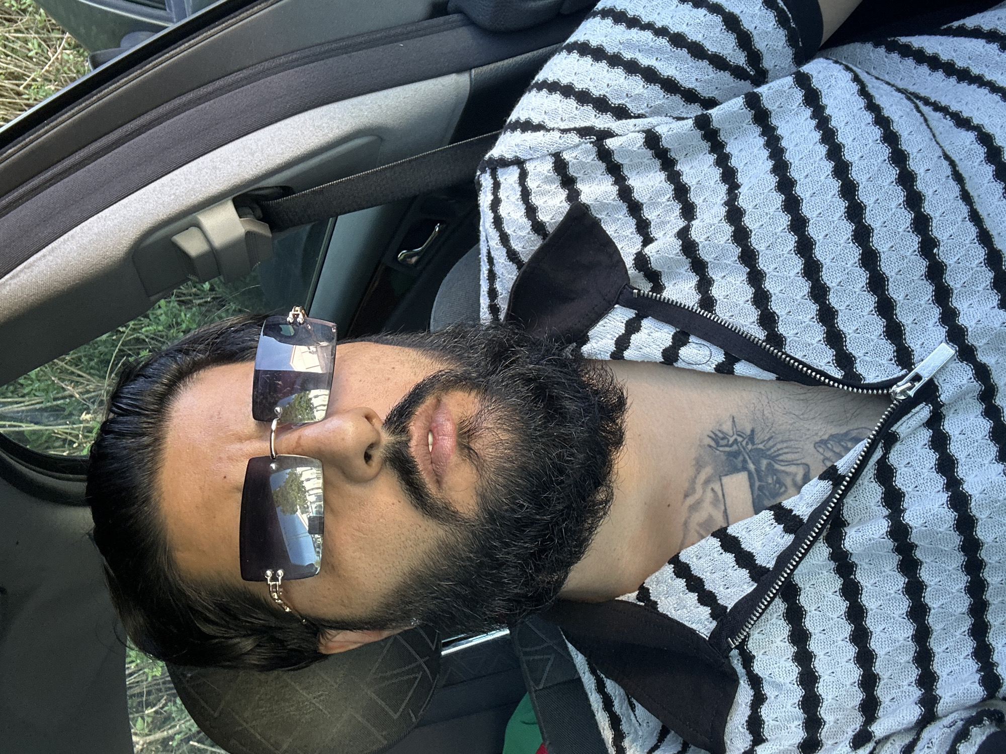 A close-up, selfie-style shot shows a person from the chest up, seated in the driver's side of a car. The person has dark hair, a full beard, and is wearing rimless, rectangular sunglasses with a gold accent. They are also wearing a black and white striped knit shirt with a zippered collar. A tattoo of Jesus with a crown of thorns is visible on the person's chest. The background shows the interior of the car, including the seatbelt and door handle, and out the window, foliage and what appears to be a building can be seen. The scene is outdoors during the day.