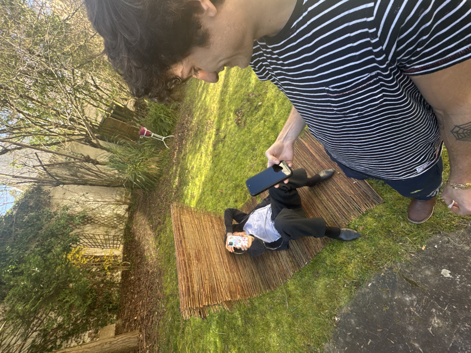 The scene is set in a sunlit residential garden in Vincennes, France. In the foreground, a male with dark, curly hair, wearing a black and white striped t-shirt, dark shorts, and brown shoes, is positioned with his right side partially visible. His right arm is extended, holding a black smartphone horizontally, aimed towards another individual. A gold-colored chain is visible on his left wrist, and a spiderweb tattoo is present on his right forearm. He exhibits a slight smile.
In the mid-ground, a second male is lying supine on a reddish-brown woven mat, which appears to be made of bamboo or reeds and is laid out on green grass. This individual is dressed in a dark jacket or hoodie over a light-colored shirt, dark pants, and dark shoes. He holds a white-cased smartphone directly in front of his face, obscuring its details.
The background consists of a tall concrete wall extensively covered with green ivy and other foliage. Various trees and shrubs, some with sparse leaves and others bare, are visible. A white metal plant stand holding a magenta pot is situated on the grass in the upper-right background. The ground is primarily green grass, with patches of exposed earth and a segment of paved or concrete surface in the lower-left foreground. The area is illuminated by bright sunlight, creating visible shadows.