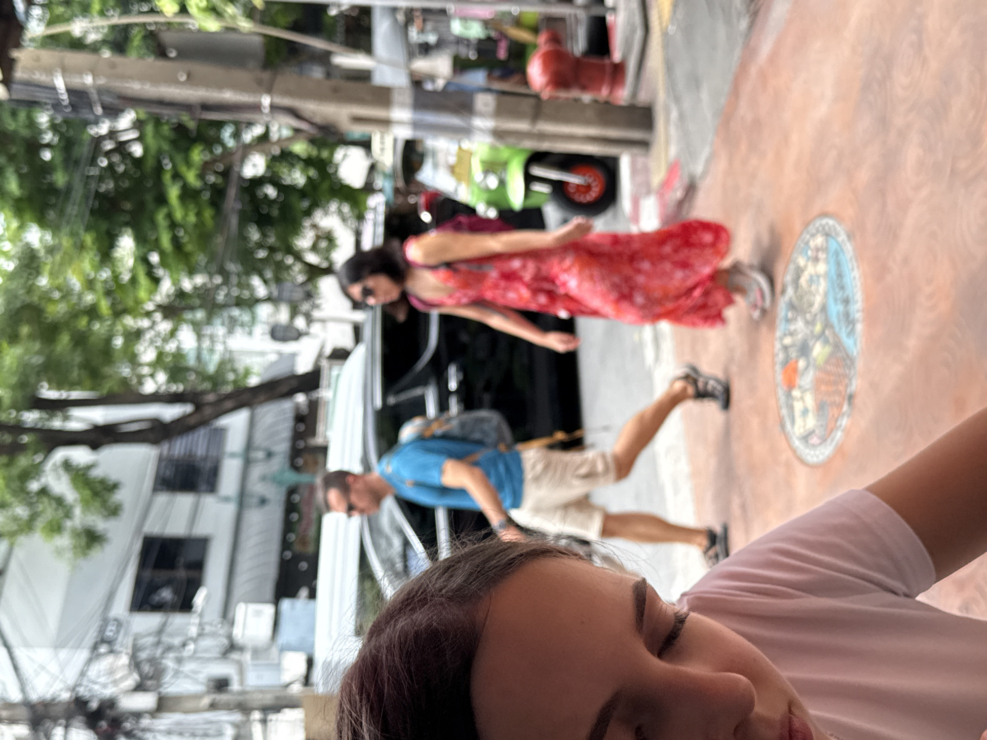 In Bangkok, Thailand, a close-up shot shows the top of a young woman's head and shoulder in the foreground, partially obscuring a street scene. She is wearing a pink t-shirt. In the background, a man in a blue t-shirt and tan shorts walks past a black SUV, wearing a backpack. To his right, a woman in a flowing red floral dress with a pink backpack walks forward, wearing sunglasses. A bright green tuk-tuk is visible in the middle ground, parked on the street, along with a red fire hydrant. The environment appears to be an urban street with trees, buildings, and overhead wires. A decorative circular inlay is visible on the sidewalk in the lower portion of the image.