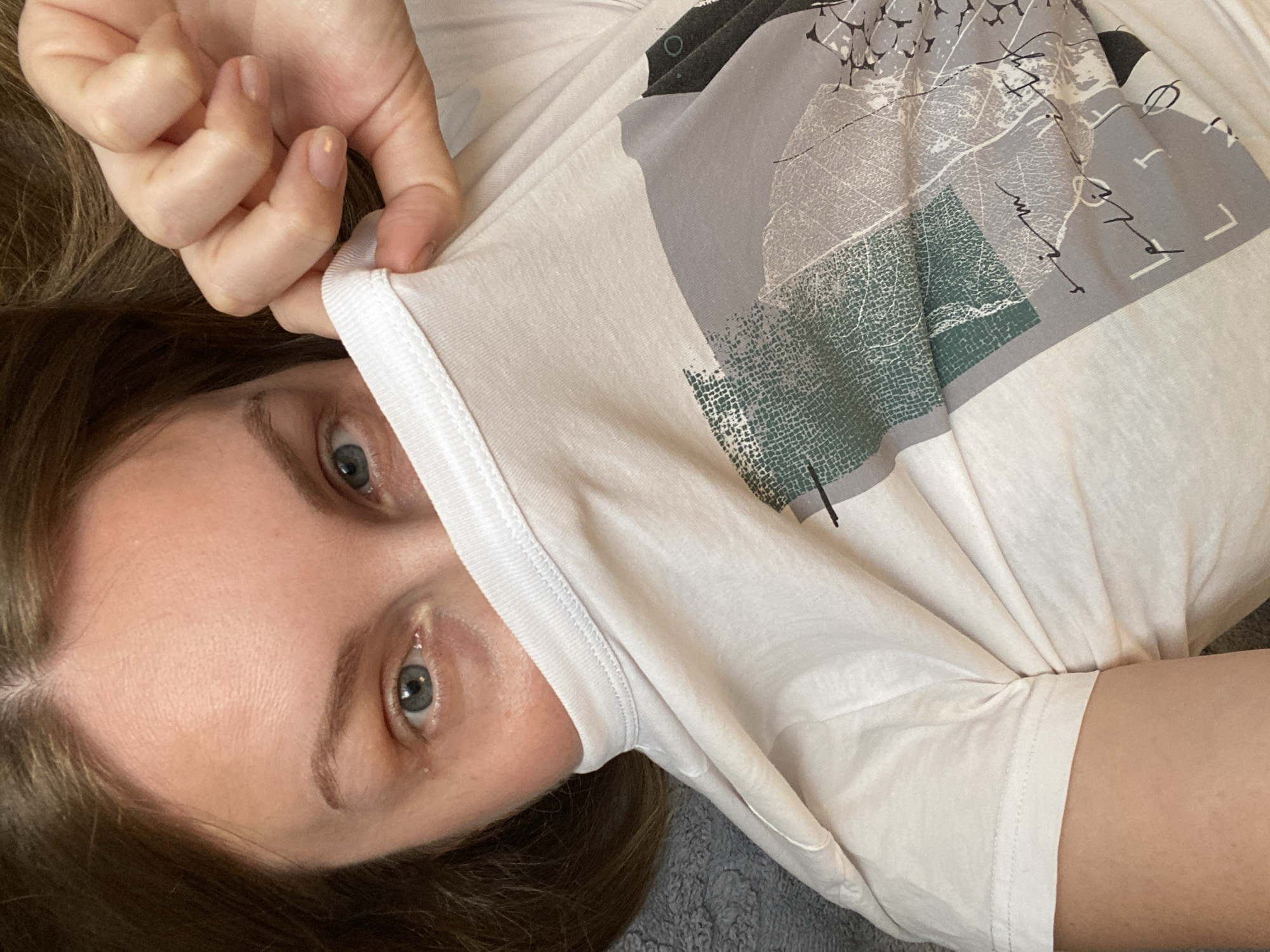 A woman with fair skin and blue eyes is positioned in the frame, partially obscured by the white t-shirt she is holding up. The t-shirt features a graphic design in shades of grey and green. Her right hand is visible holding the fabric of the t-shirt. The background is a blurred, neutral tone.