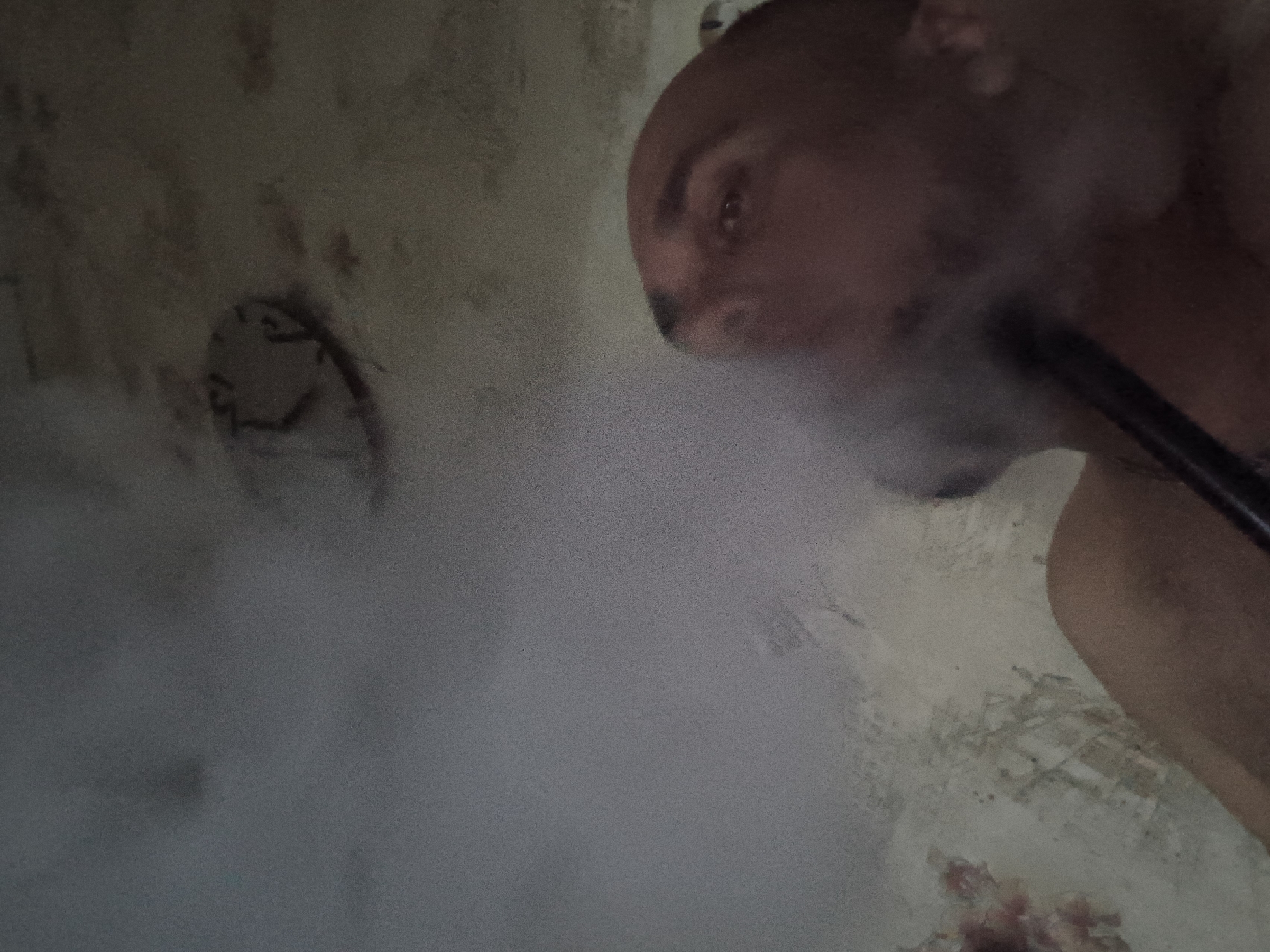 The image depicts a close-up, low-angle view of a person exhaling a cloud of smoke, obscuring much of the scene. The subject appears to be a bald individual with dark eyebrows, and a portion of their face and chest is visible. They are holding a dark object to their mouth, from which the smoke is emanating. The background shows a textured, dimly lit ceiling with what appears to be an old, circular fixture or marking on it. The wall beneath the ceiling has a mottled, aged appearance with some reddish-brown stains and what may be faded markings or repairs. The lighting is subdued, creating a hazy and somewhat claustrophobic atmosphere. There is no discernible information within the image to associate it with the city of Zuevka, Russia, or any specific location.