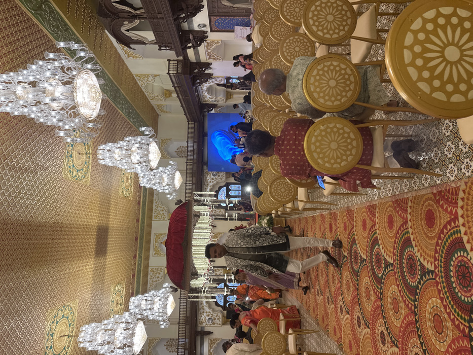 A large hall features rows of seated people, many observing a figure walking under a red canopy. Elaborate chandeliers hang from the ceiling, illuminating the golden walls. A sizable screen displaying blue graphics is visible in the background. The floor is covered with patterned carpets, and intricately designed chairs provide seating. Balconies with ornate wooden carvings overlook the hall.