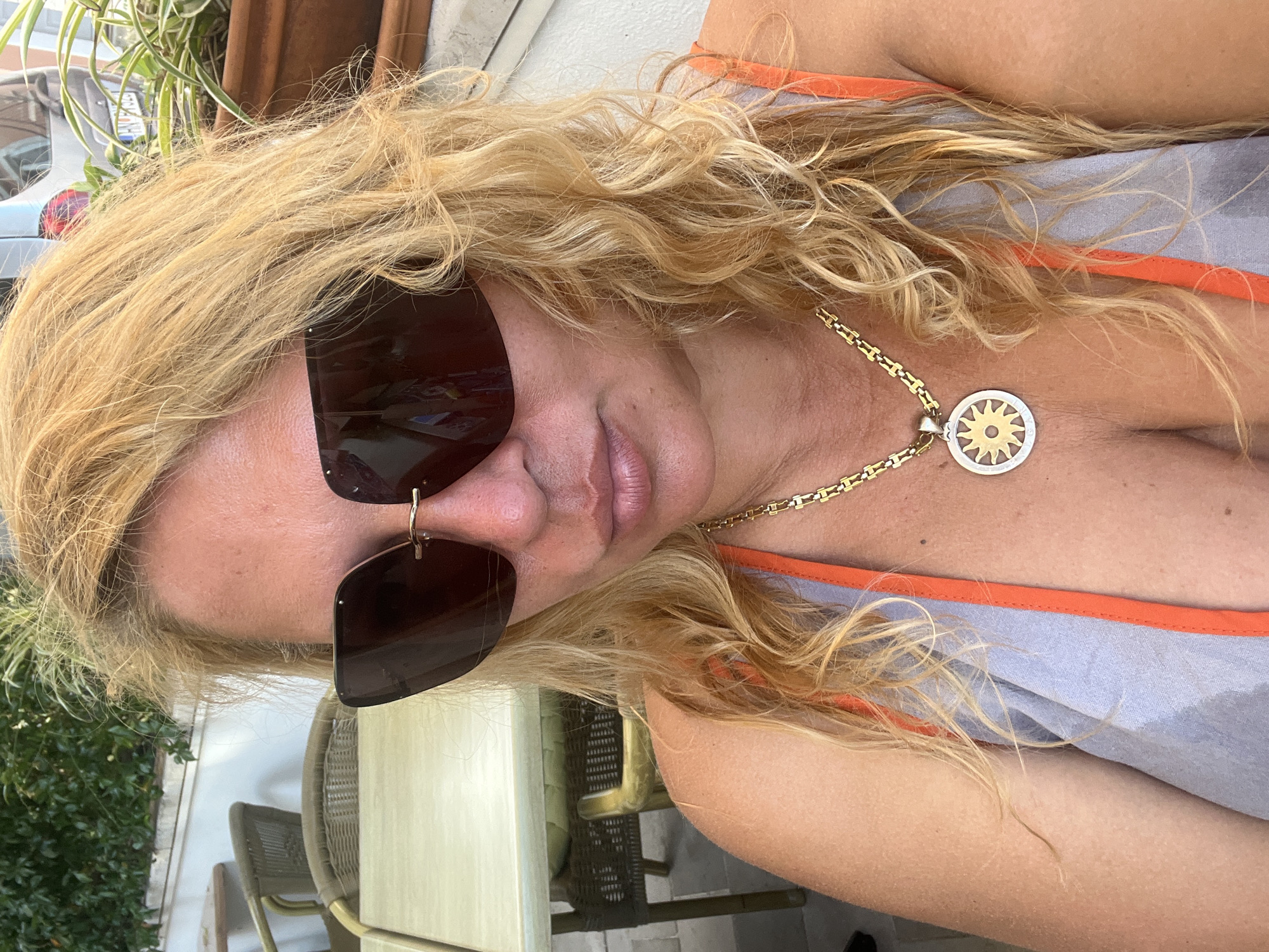 A close-up, over-the-shoulder selfie taken outdoors shows a blonde woman with curly hair wearing large dark sunglasses, an orange and gray tank top, and a gold chain with a sun pendant. She is seated at an outdoor cafe with tables and chairs visible in the background. The environment suggests a warm climate, possibly a seaside town. The location is Đenovići, Montenegro.