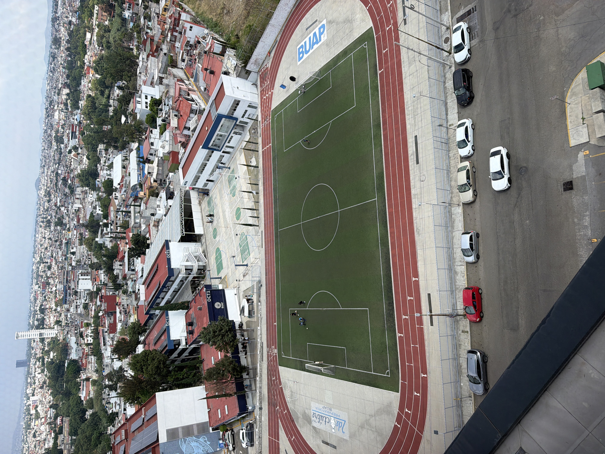 The image shows an aerial view of a BUAP (Benemérita Universidad Autónoma de Puebla) Preparatoria stadium and athletic field in San Bernardino Tlaxcalancingo, Mexico. The stadium includes a running track surrounding a green soccer field. The soccer field has standard markings, including the center circle and goal areas. On the field, there are a few individuals who appear to be in the middle of a training session. The stadium has a concrete walkway and a light-colored building adjacent to the field. There are several parked cars near the stadium. Beyond the athletic field and building, there's a dense urban landscape with houses, buildings, and trees. The sky is overcast and the overall view is looking down at a significant angle.