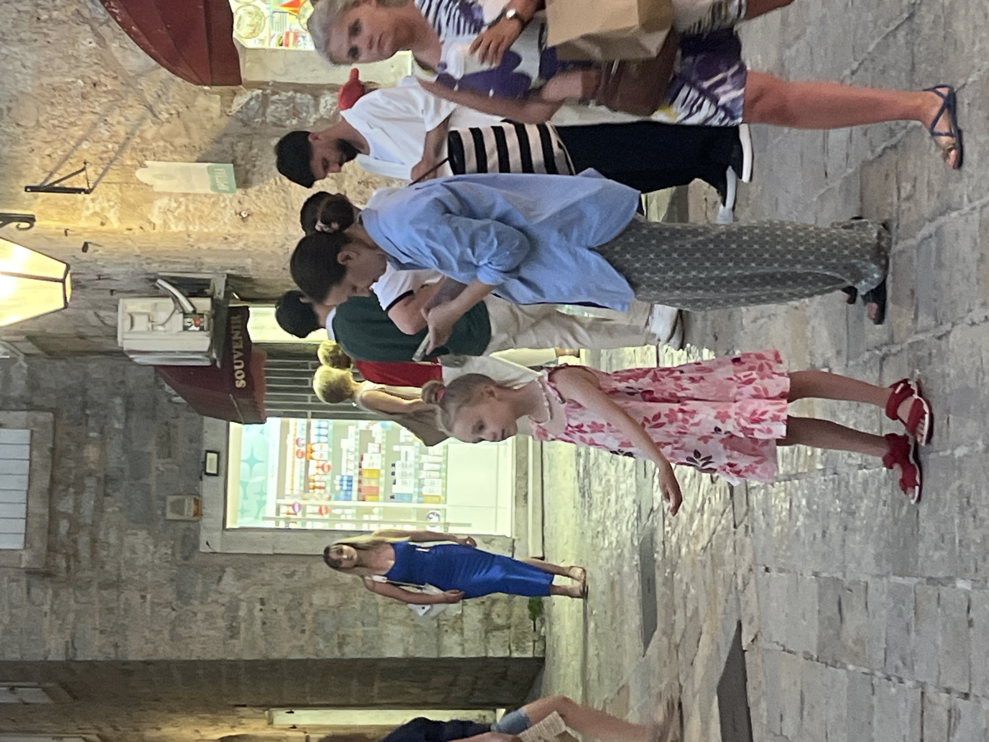 In Budva, Montenegro, on a cobblestone street, a young girl in a pink floral dress and red sandals stands facing left, her right hand outstretched. Behind her, a woman in a blue shirt and patterned pants looks at her phone, standing next to a woman with a bun and a man in a white shirt and black and white striped pants. Further back, a woman in a blue dress walks away from the camera. To the left, a pharmacy with illuminated shelves is visible. The scene is illuminated by artificial lights.