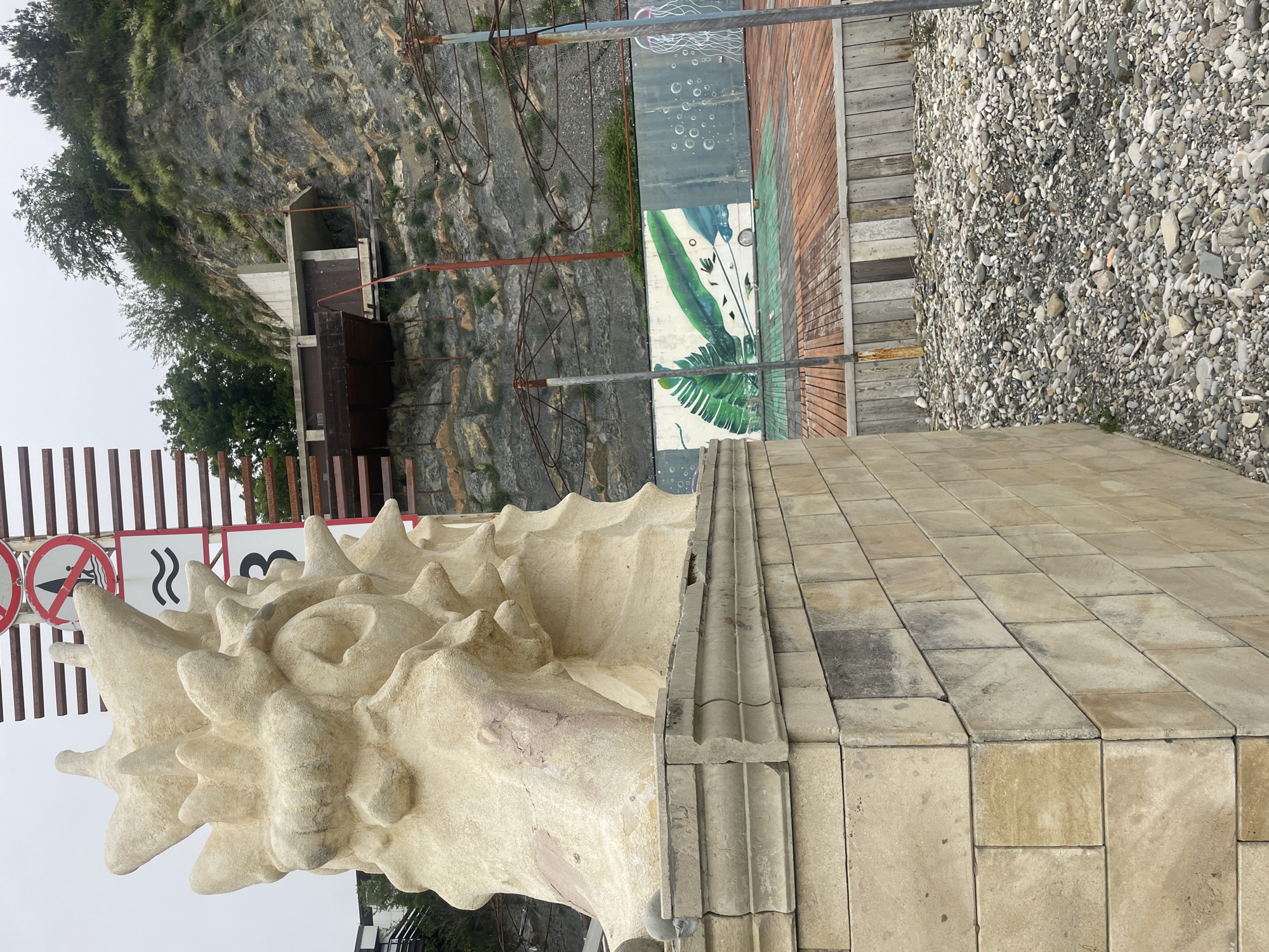 The image shows a coastal area in Olginka, Russia. A large, ornate sculpture resembling a sea shell sits on a stone base made of rectangular blocks, positioned beside a paved area. Behind the sculpture, a cliff face rises with vegetation and a concrete structure with a staircase. A metal barrier system runs along the top of the cliff. The background also features a wall with mural art, a gravel area, and signs with white and red colors. The image suggests a seaside promenade or park.