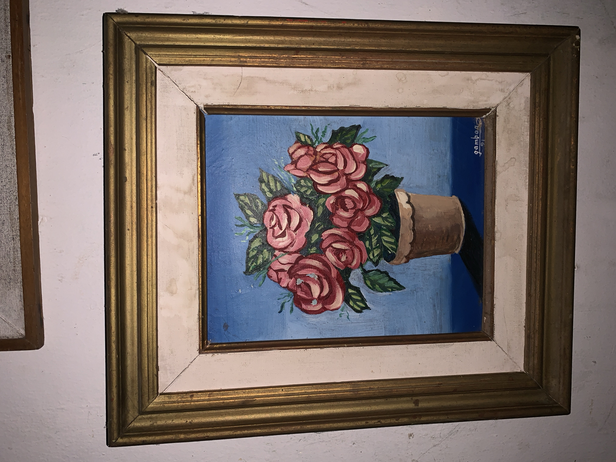 A framed painting of red roses in a brown pot is hanging on a white wall. The frame is a combination of gold and white. Another framed object is partially visible to the left. The location in Santa Ana, Venezuela is not determinable from this image alone.