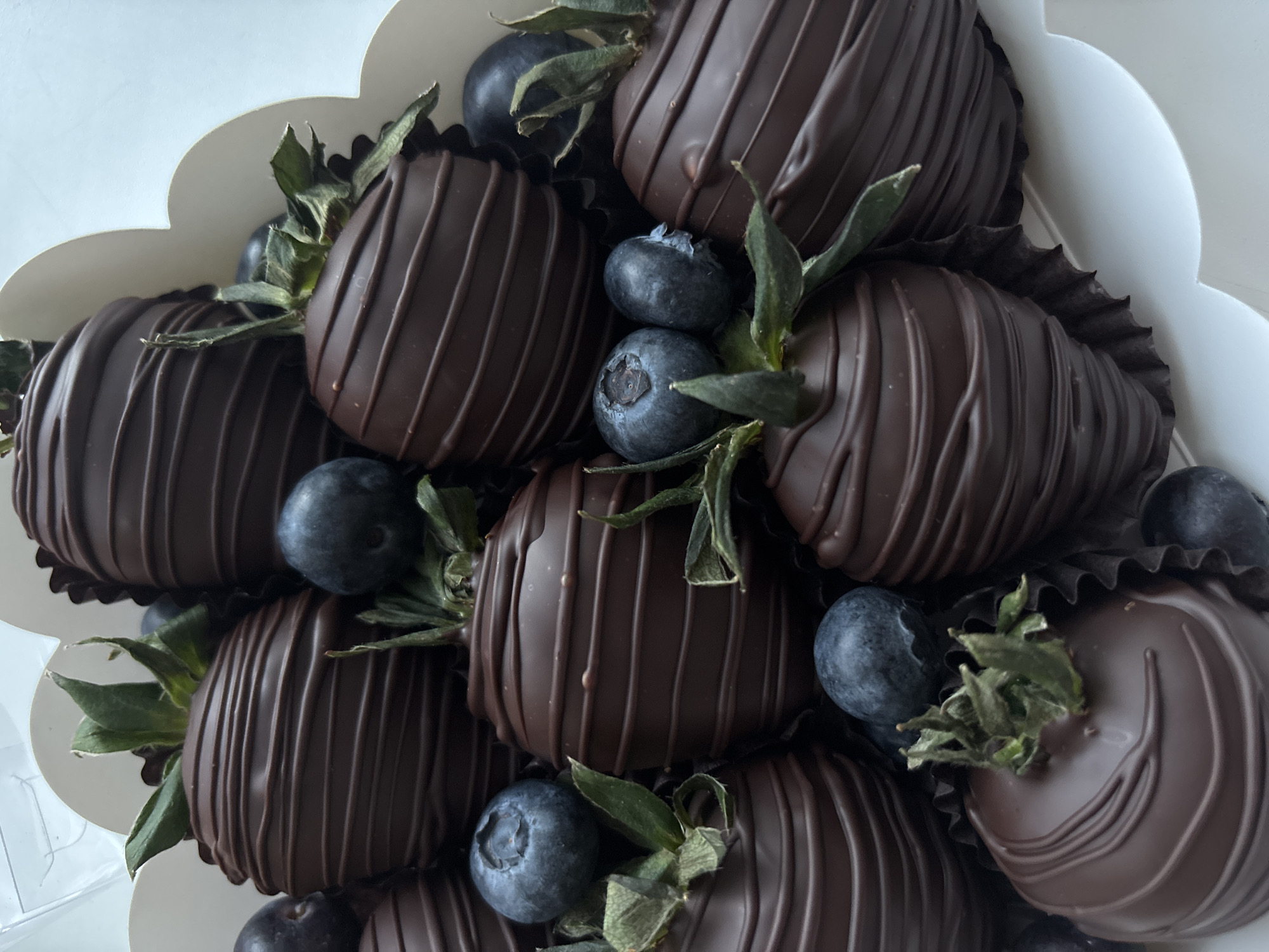 The image displays a close-up, overhead view of a confectionery arrangement primarily consisting of chocolate-covered strawberries and fresh blueberries. Approximately ten to twelve dark chocolate-enrobed strawberries are visible, each featuring thin, horizontal drizzle patterns of the same dark chocolate. The green calyx and partial stems remain attached to the top of most strawberries. Each strawberry is individually cradled in a dark, fluted paper liner. Numerous spherical, deep blue blueberries are interspersed among the strawberries. The entire arrangement is contained within a white box, the upper edge of which presents a distinctive scalloped or wavy design. A partial, transparent cover, likely plastic, is visible in the upper left background. The lighting is bright and even, indicating an indoor setting. No individuals are present, and no discernible details within the image contextually indicate the asserted location in the city of Perm, Russia.