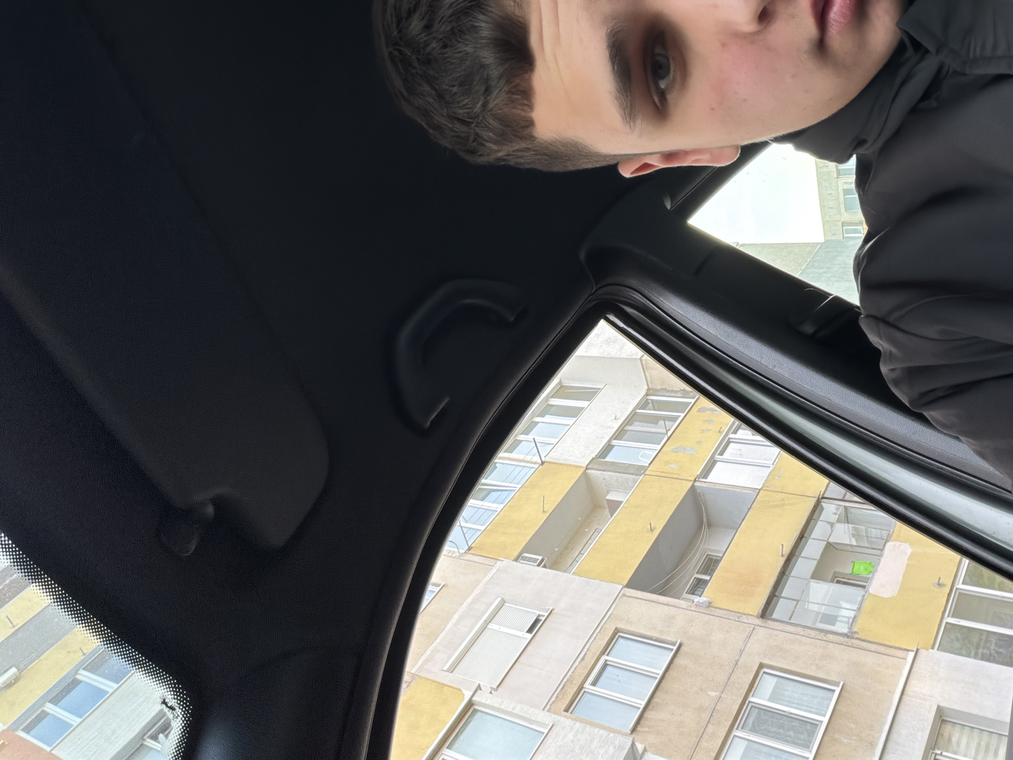 The image shows a low-angle view from inside a car, looking up at a building. A young man is partially visible in the top portion of the frame. The building is multi-storied with a patterned facade of yellow and white sections, each containing multiple windows. The car's interior includes the headliner, sun visor, and a passenger-side assist handle. The scene appears to be in Sofia, Bulgaria based on the architectural style of the building.