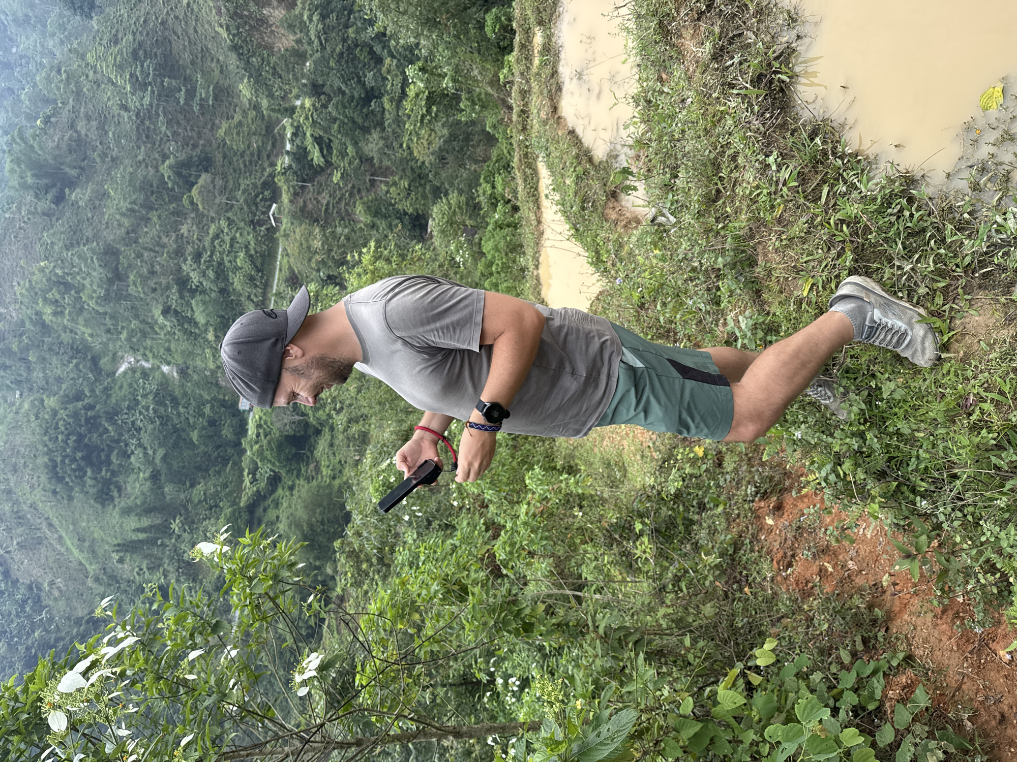 The image depicts a man standing on a dirt path next to a water-filled agricultural terrace, set against a backdrop of lush green mountains. The scene is characteristic of a mountainous, vegetated region, specifically in Nậm Dịch Commune, Vietnam.
The man, positioned on the right side of the frame, is seen in profile, looking down. He wears a grey baseball cap, a plain grey t-shirt, green and black shorts, and grey sneakers. Accessories include a black smartwatch on his left wrist and red and blue bracelets on his right wrist. He holds a black rectangular electronic device, possibly a smartphone or small camera, which is connected by a red cord. His posture suggests he is focused on the device in his hands.
To the man's right, a muddy, shallow body of water, likely part of a terraced rice paddy system, extends along the edge of the path. The banks of this water feature are overgrown with various green plants and grasses, and small dark specks, possibly insects, are visible on the water's surface. In the background, dense, verdant mountains covered in trees and foliage rise, appearing somewhat hazy in the distance. The foreground also features various green plants and bushes.
The time of day appears to be daytime, possibly overcast or with soft light, as strong shadows are absent. The overall impression is one of a remote, natural environment with agricultural elements. There is no visible text in the image.