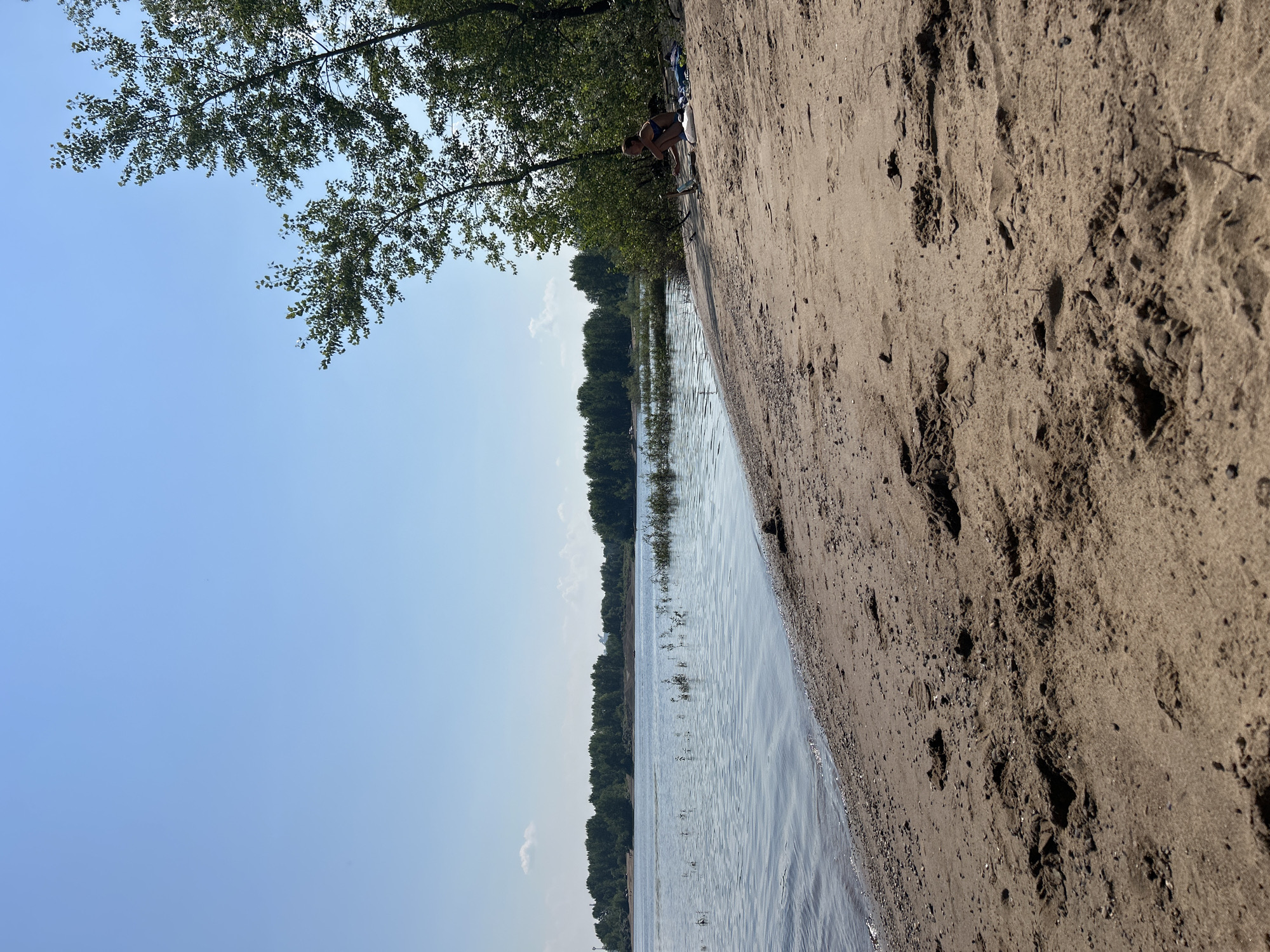 The image depicts a natural scene on the sandy shore of a wide river, likely the Volga River, in Noviy, Russia. The foreground is dominated by wet sand with visible footprints, leading down to the water's edge. The river itself is calm, with ripples reflecting the clear blue sky. Along the far bank, a dense line of trees forms a natural horizon. To the right, on the sand near the treeline, a young woman is sitting with her back to the camera, wearing a dark bikini. She appears to be interacting with an object on the sand in front of her, possibly a phone or book. Several items, including a blue towel, are placed next to her. The overall environment is sunny and peaceful, suggesting a recreational setting.