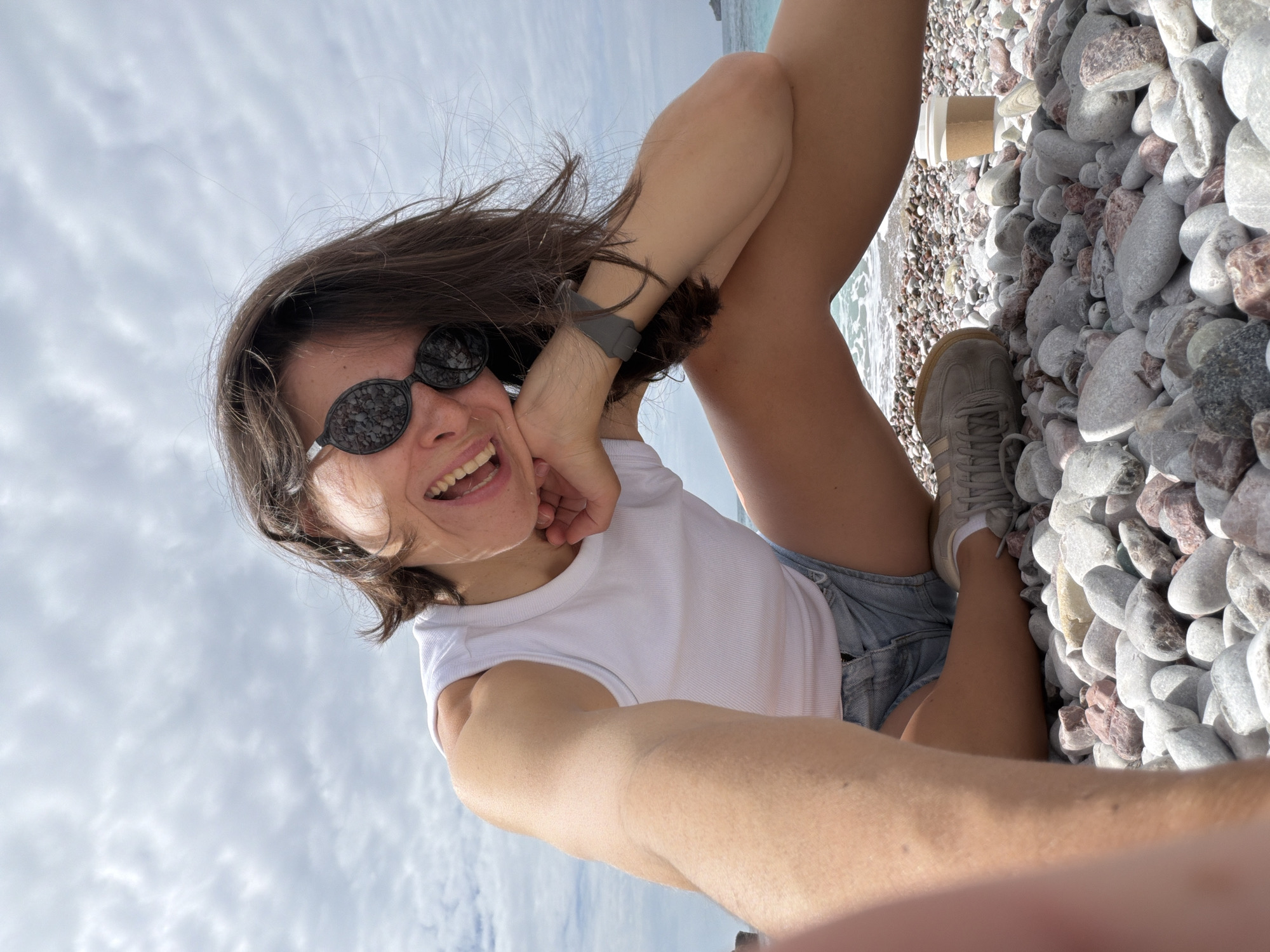 The image shows a woman in Rafailovići, Montenegro, lying on a pebble beach. She is wearing sunglasses, a white sleeveless top, and denim shorts. One leg is raised, and she's holding her head with one hand, smiling. She wears a grey smartwatch on her left wrist. One grey sneaker is visible near a white paper cup placed on the pebbles. The background features the sky with clouds. The beach stretches towards the water, which is visible in the top right corner of the image.