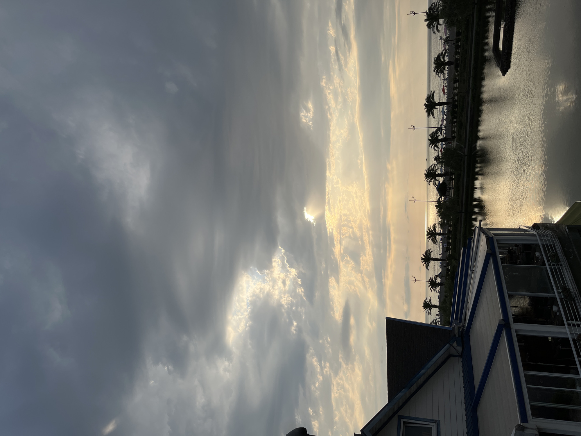 A view from a building shows a cloudy sky with some sunlight breaking through. In the foreground, a building with a white and blue facade and a dark roof is visible. Beyond the building, there is a body of water reflecting the sky and palm trees. Across the water, there is a beach with several palm trees, beach umbrellas, and people. The sea is visible in the background, with a faint horizon line. The image appears to be taken in Batumi, Georgia, given the characteristic architecture and palm trees.