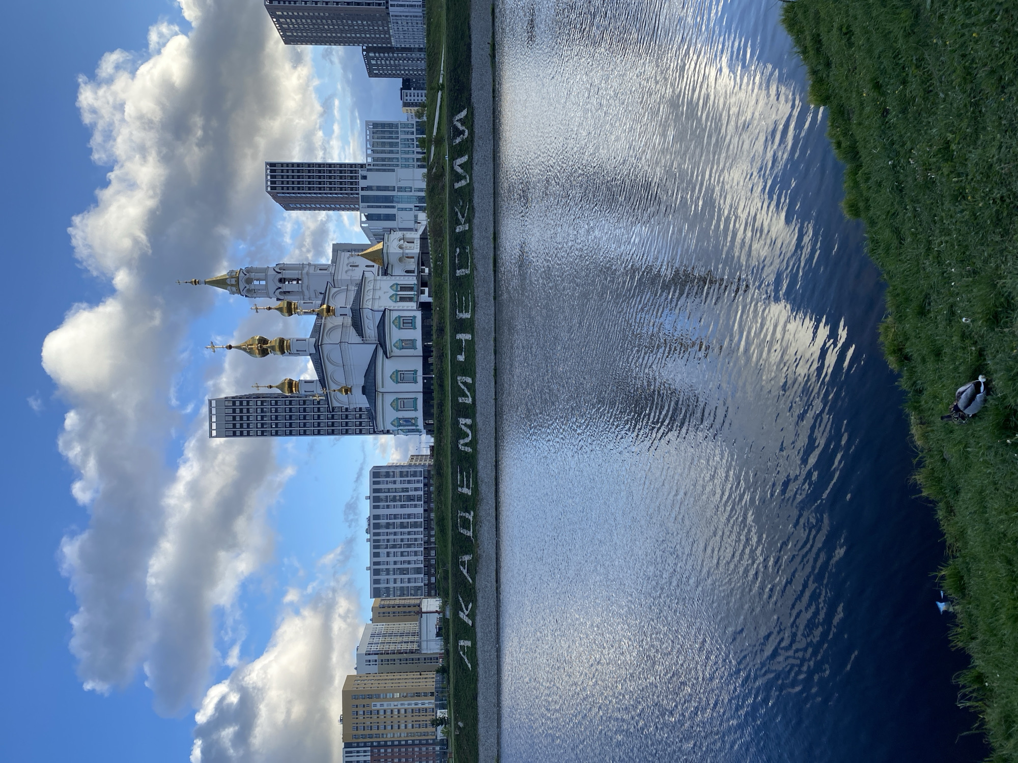 The image is taken in Yekaterinburg, Russia, at a canal or pond with the word "AKADEMICHESKII" spelled out in landscaping along the bank. In the background is the Church on the Blood in Honor of All Saints Resplendent in the Russian Land, a modern Russian Orthodox church. Also in the background are several high-rise apartment buildings. The sky is blue with scattered clouds. The canal's surface is rippled with reflections of the sky. A duck is visible at the lower right corner of the image, on the grassy bank.